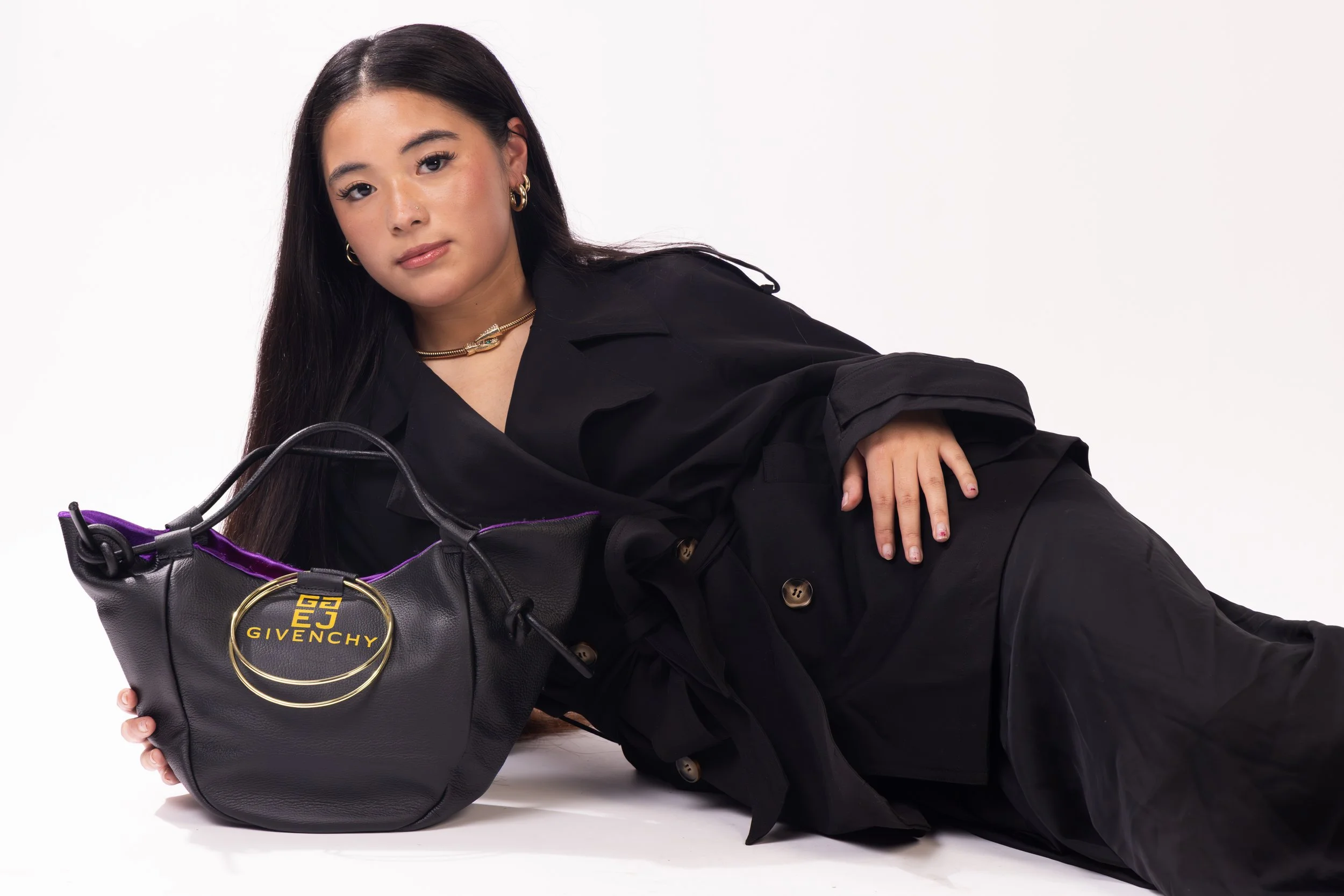 A woman with long dark hair wearing black clothing, lying on her side with a black Givenchy handbag with gold hardware, posing against a plain white background.