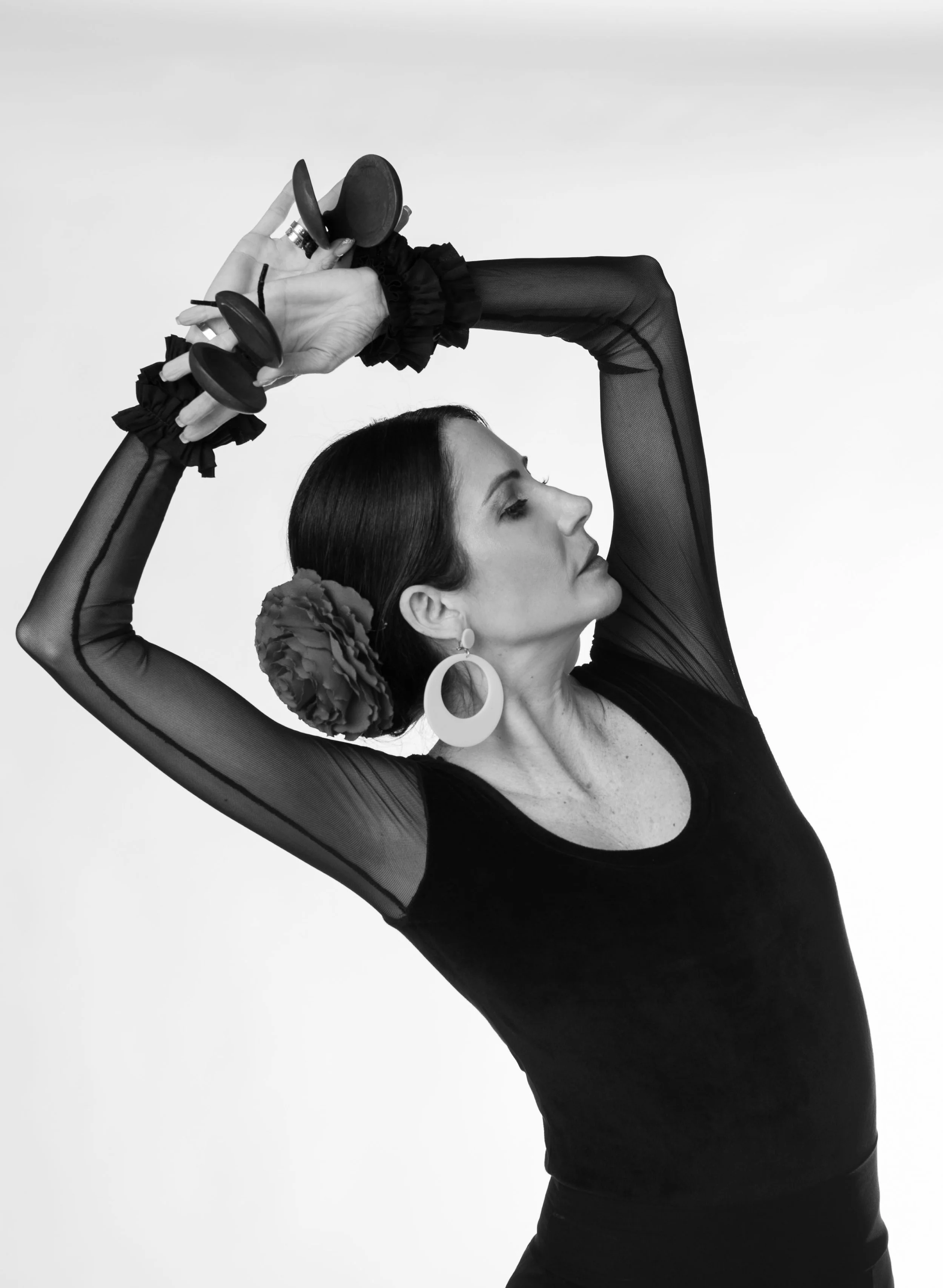 Black and white photo of a woman with a flower in her hair, wearing large hoop earrings and a black top with mesh sleeves, posing with arms raised and holding castanets.