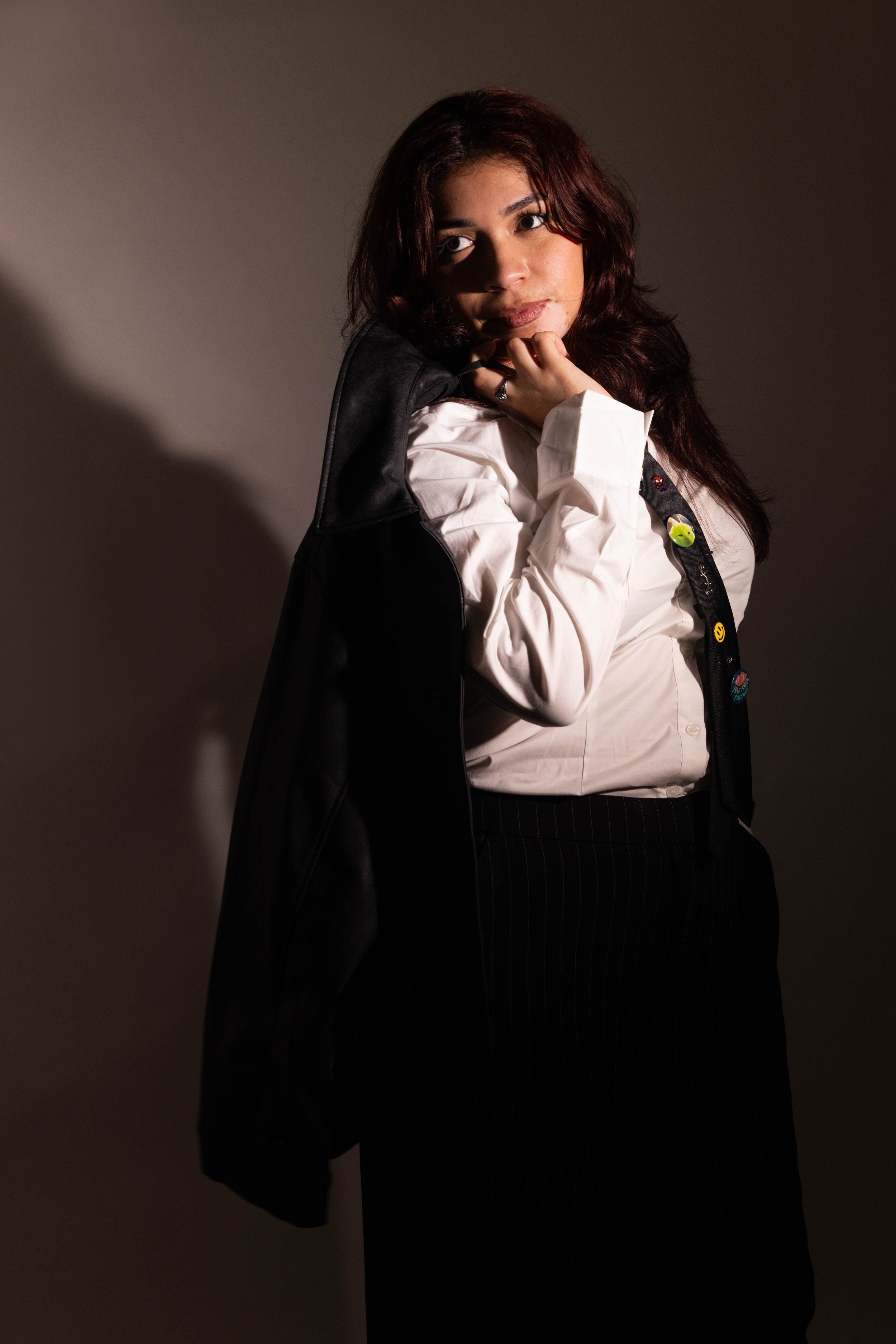 A woman with dark red hair wearing a black leather jacket, a white shirt, and a black tie with colorful patches, standing against a plain background.