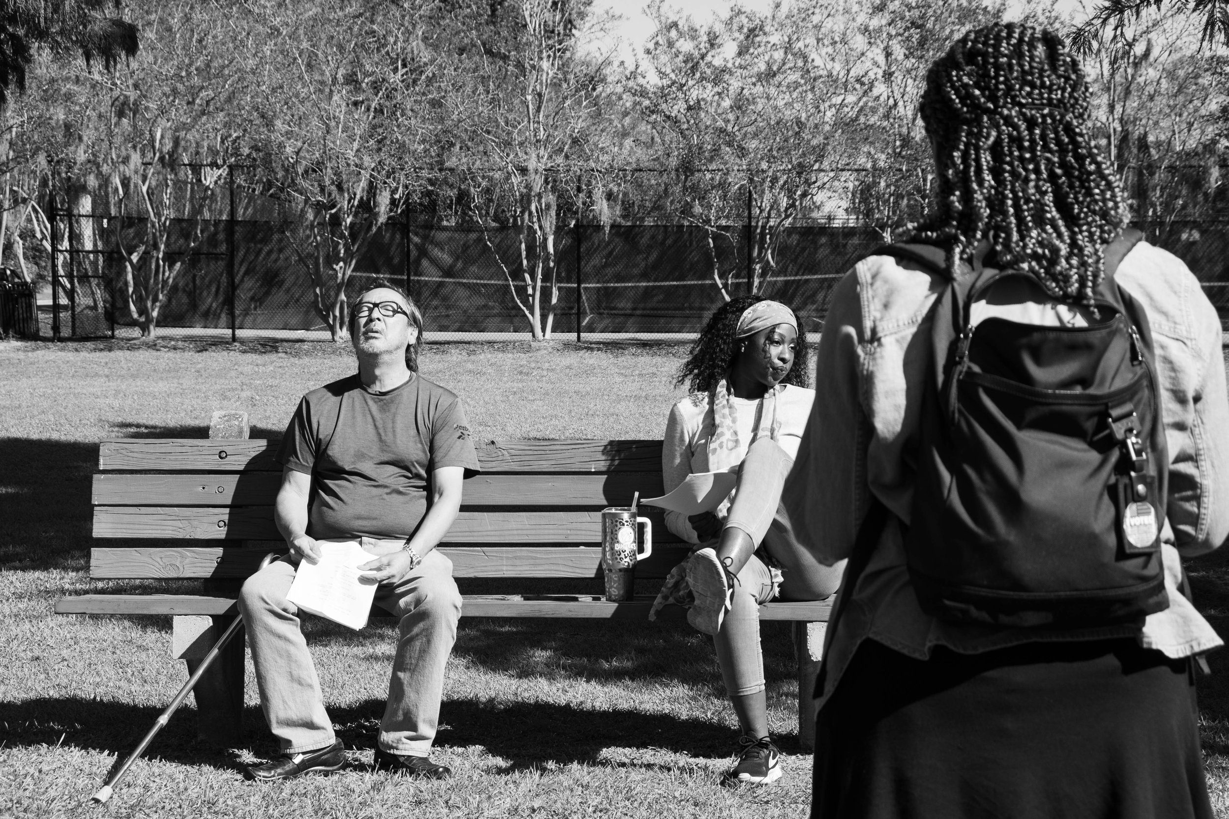 Three people outside in a park: one man sitting on a bench with a cane, a woman sitting on the bench with papers and a drink, and a woman with braided hair standing in front of them with a backpack.