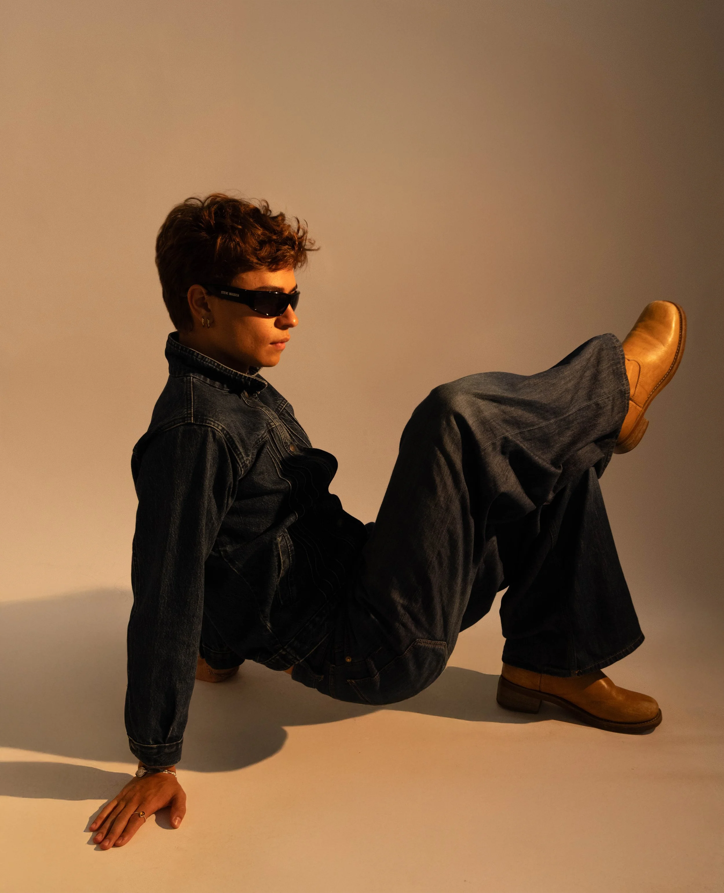 Person sitting on the floor, wearing sunglasses, a denim jacket, dark jeans, and brown boots, with legs raised and one hand on the ground.