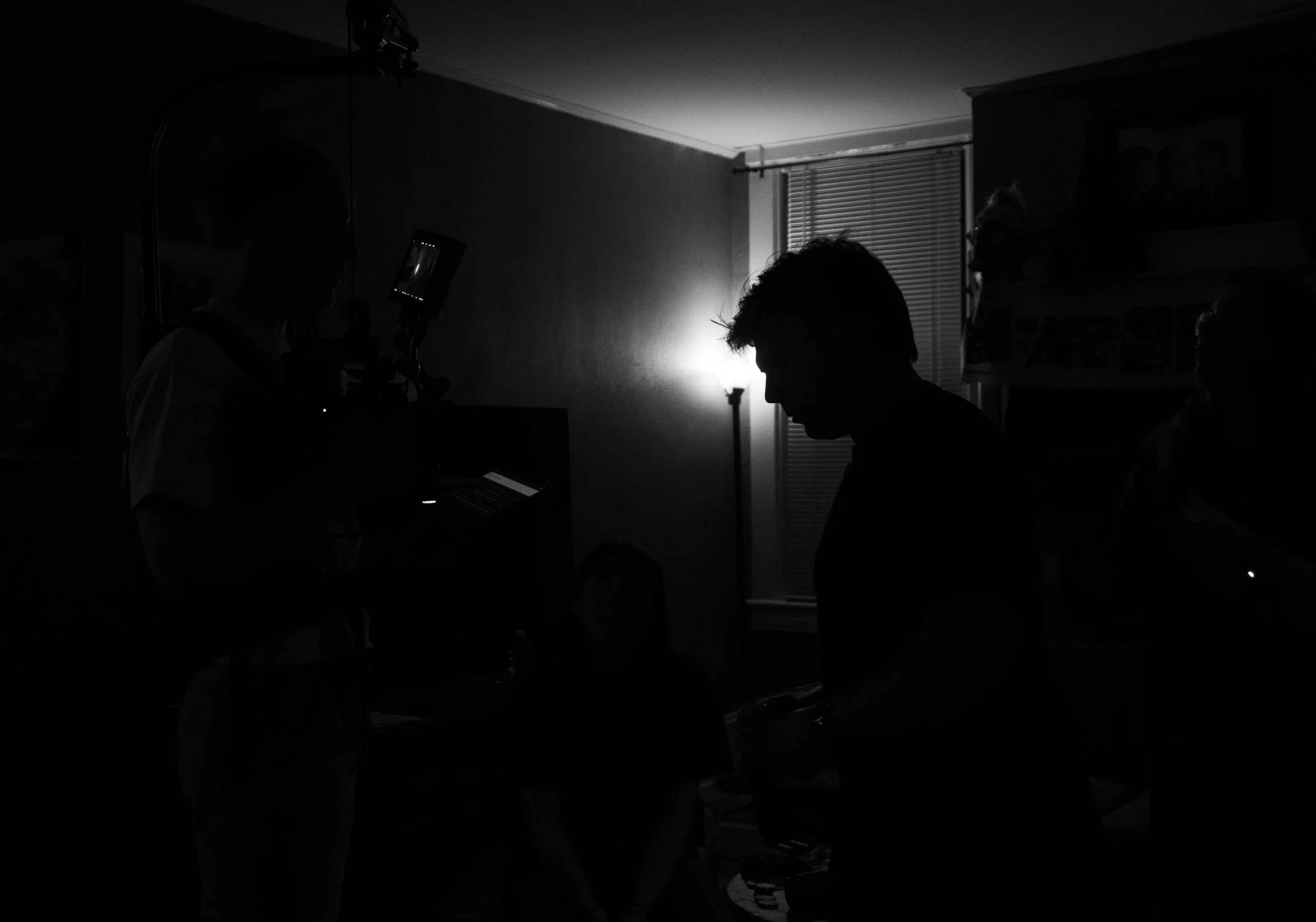 A silhouette of people in a dimly lit room, with some individuals holding equipment and others standing or sitting, illuminated by a single light source from a window and a lamp.