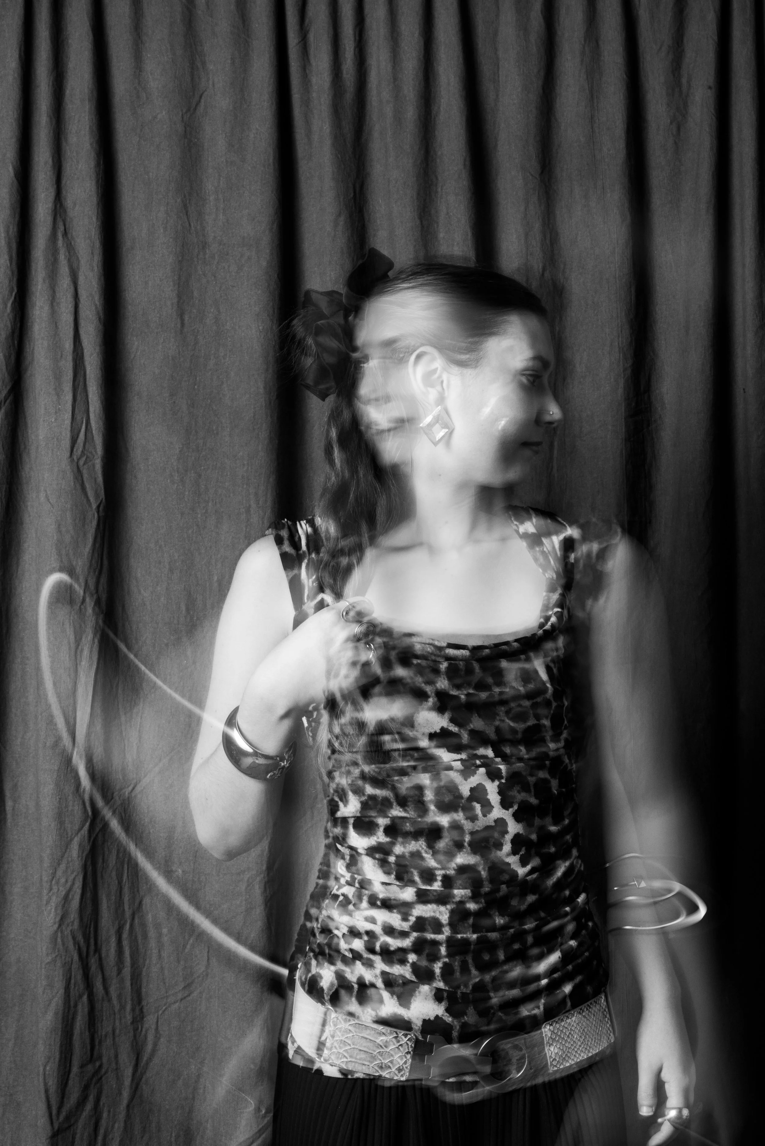 A woman wearing a leopard print top and jewelry, standing in front of a dark curtain, with her motion-blurred figure appearing as a double exposure.