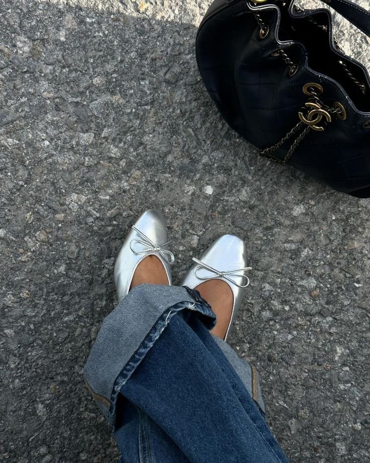 Person wearing white ballet flats and rolled-up jeans, sitting on a rough asphalt surface next to a black quilted Chanel handbag with gold chain and logo.