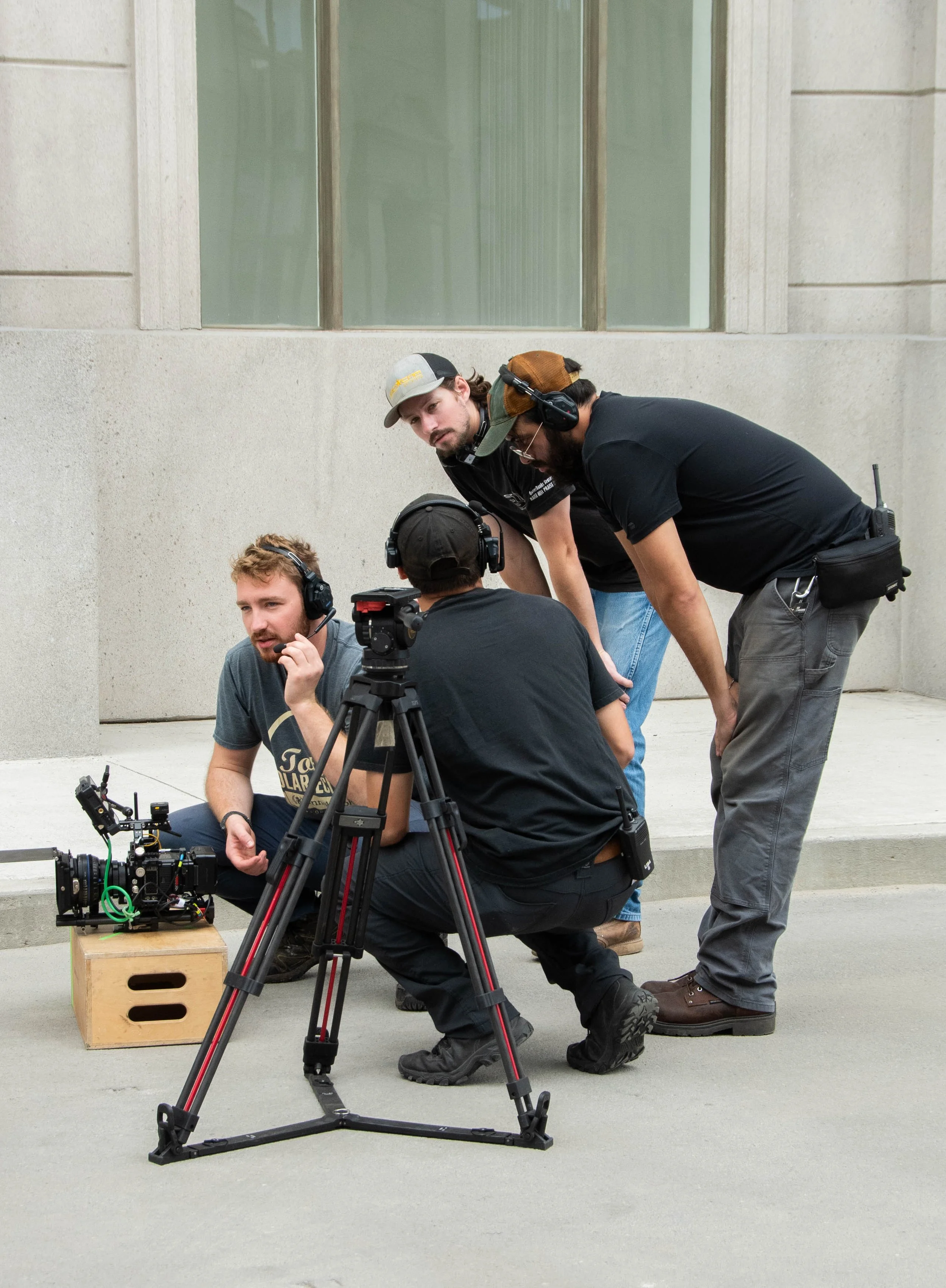 Four men with professional filming equipment on a city sidewalk, some wearing headphones and one sitting with a camera while others stand and observe.