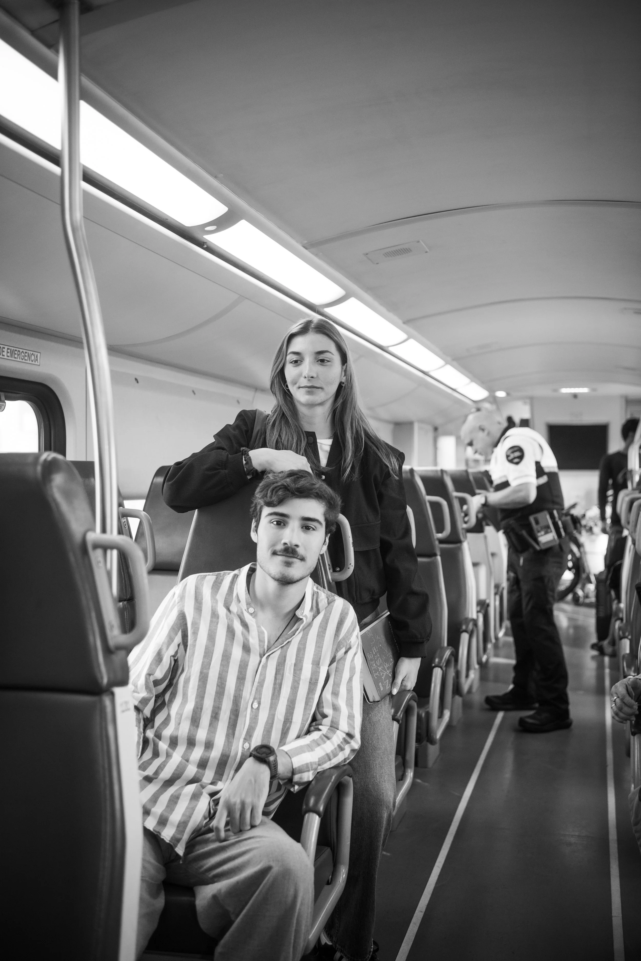 A young man with a mustache and beard in a striped shirt seated on a bus, with a young woman standing behind him with her hand on his head. Police visible in the background.