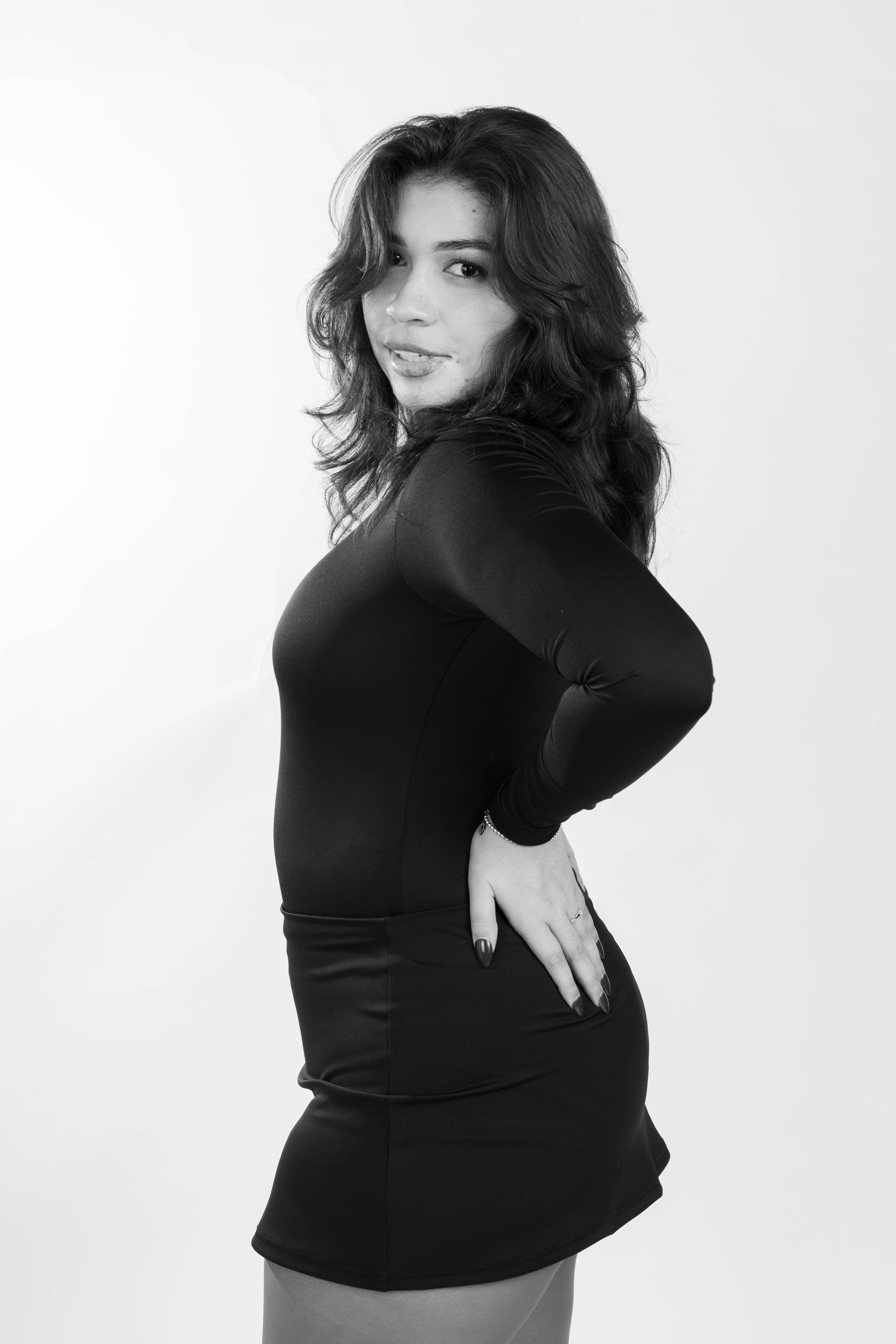 A woman with wavy hair wearing a long-sleeve black dress, standing in profile with one hand on her hip, smiling softly, against a plain background.