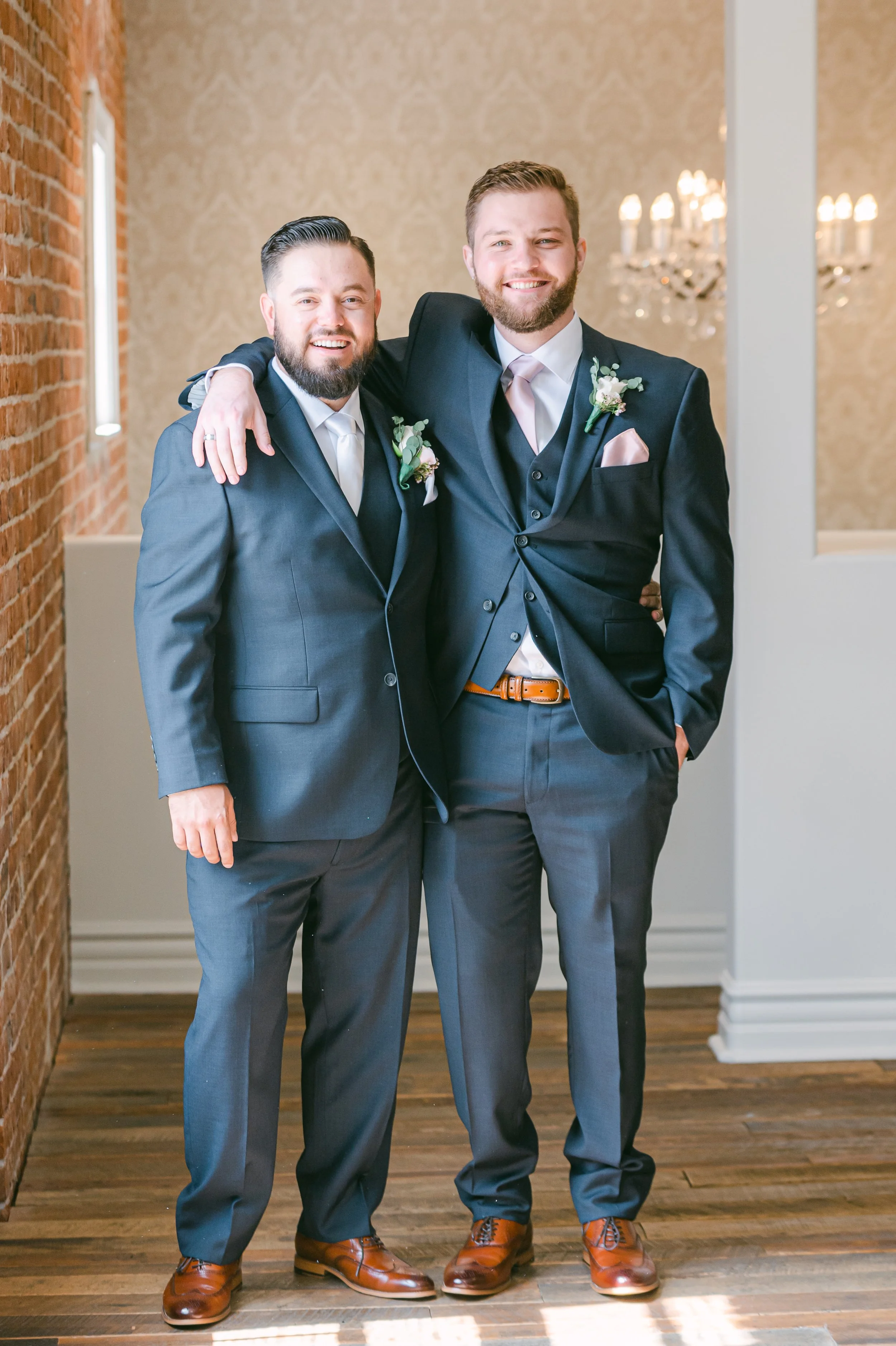 Two men in suits smiling, one has arm around the other, standing inside a room with wooden floors, brick wall on the left, decorated wallpaper on the back wall, and a chandelier in the background.