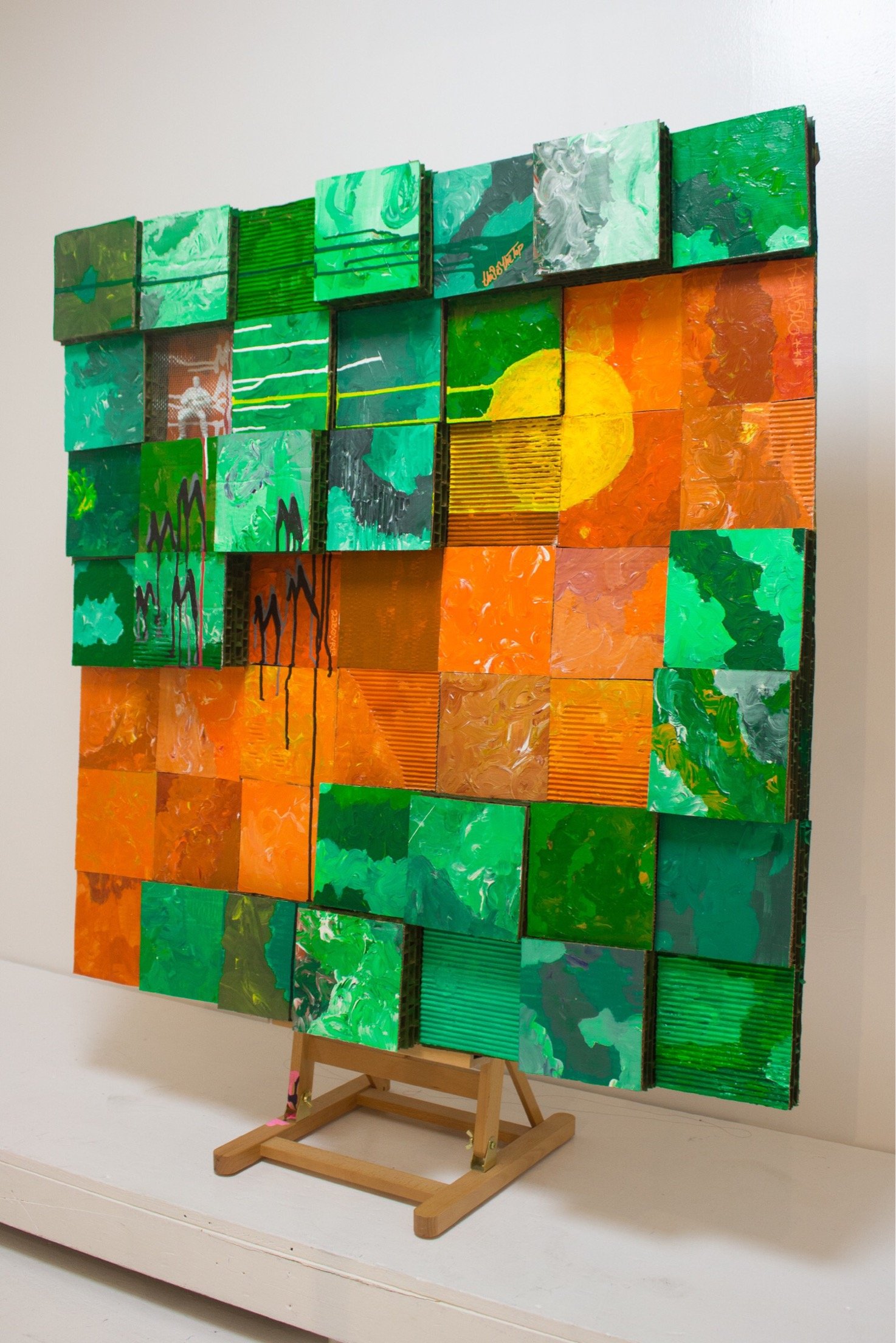 A mixed-media art piece composed of cardboard squares arranged in an abstract design. Green squares dominate the top and bottom half of the piece with the squares rising at differing levels for depth. An orange river of squares run through the center