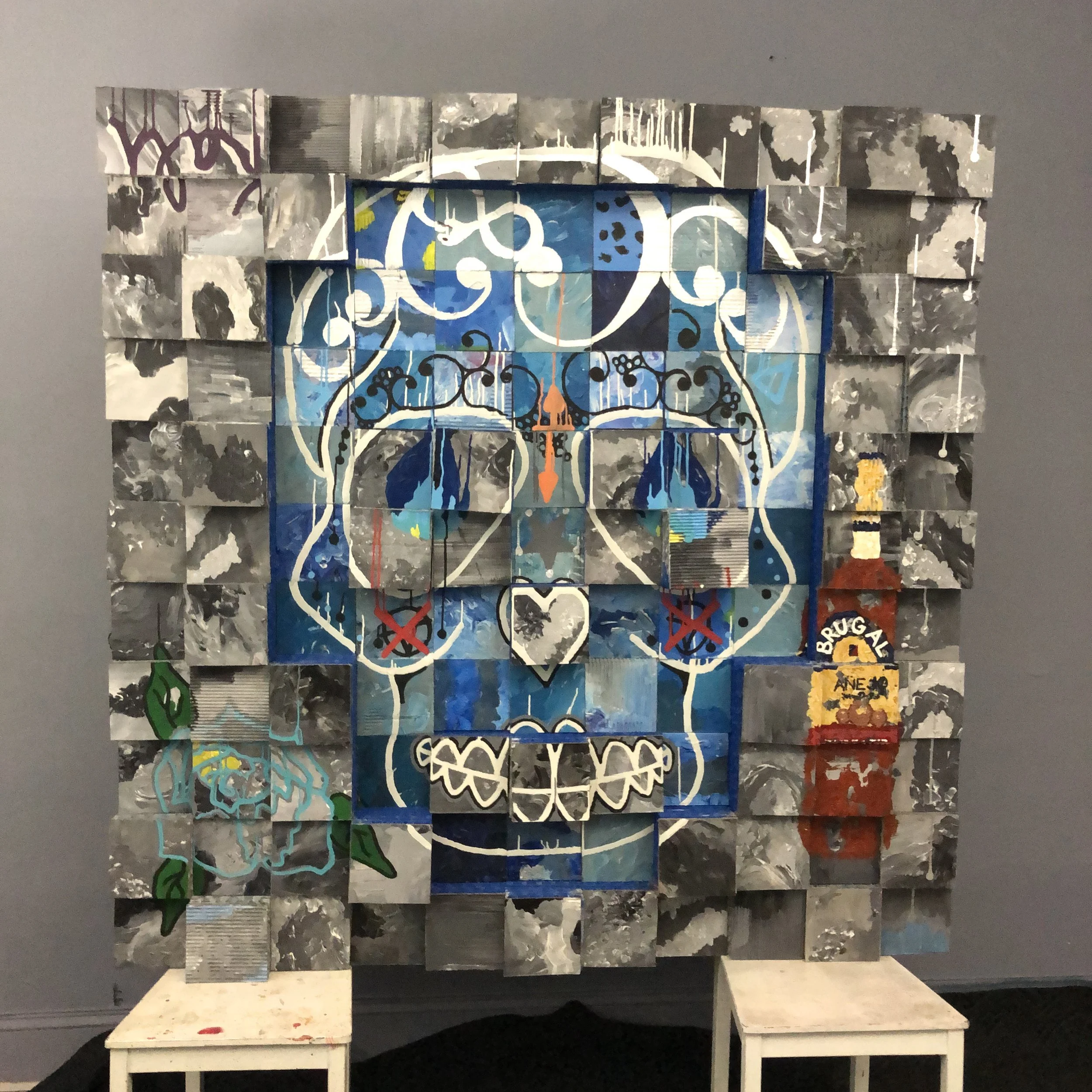 A mixed-media, grid-like art piece composed of cardboard squares arranged in an abstract design, with a blue colored skull in the center surrounded by gray. With a bottle of "BRUGAL" painted on the lower right section and a flower on the left..