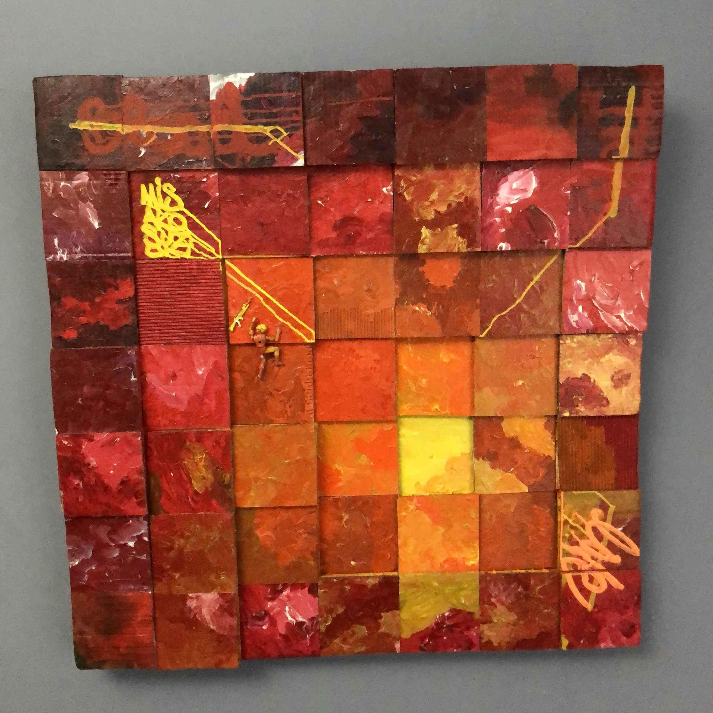 A large grid using cardboard painted squares, featuring various shades of red eventually going from orange to a single yellow square in the center of the piece. The squares are at their most stacked by the edges and the yellow center is the lowest.