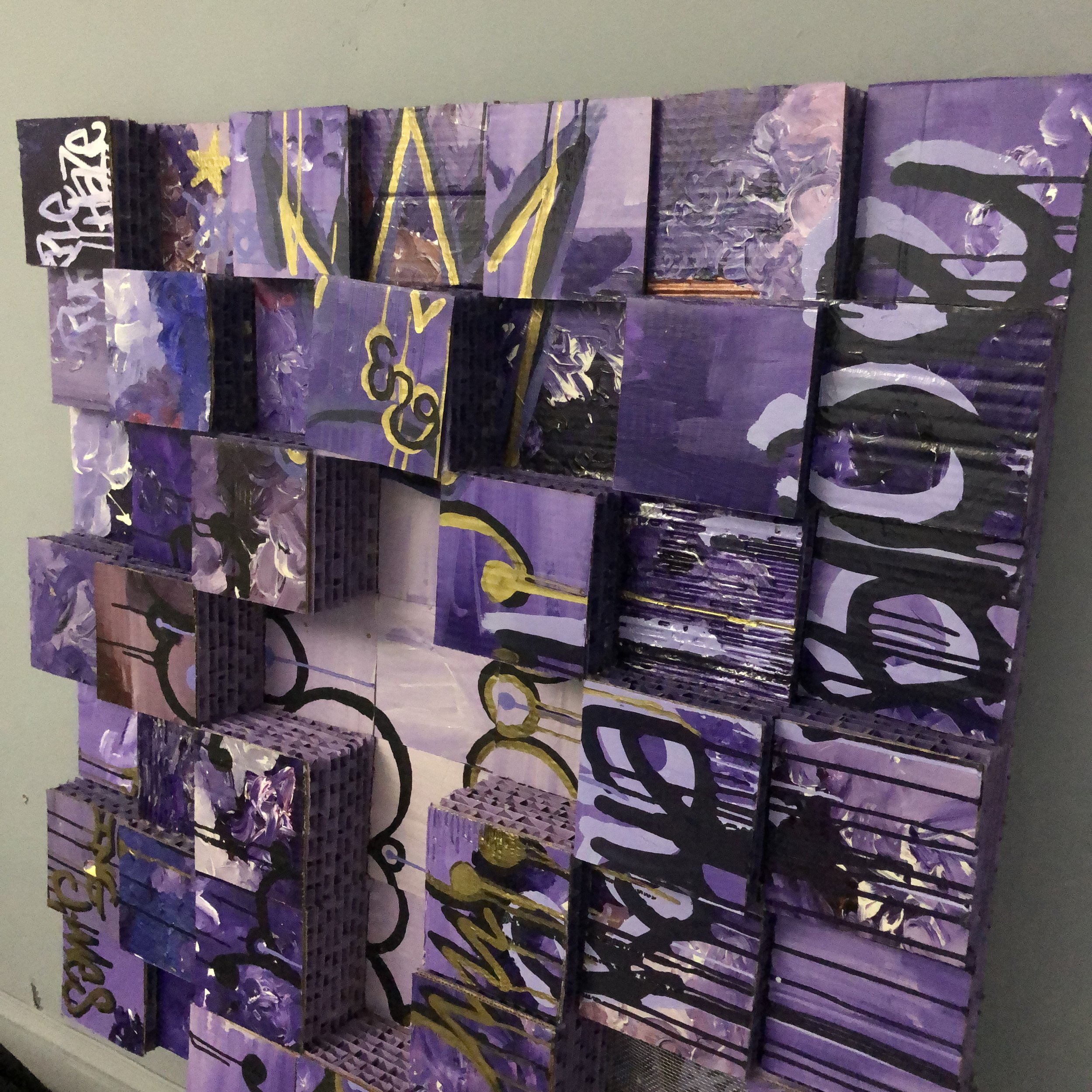 A multi-layered art installation made of painted cardboard boxes, featuring purple, black, white, and gold colors with abstract and graffiti-style designs and text.