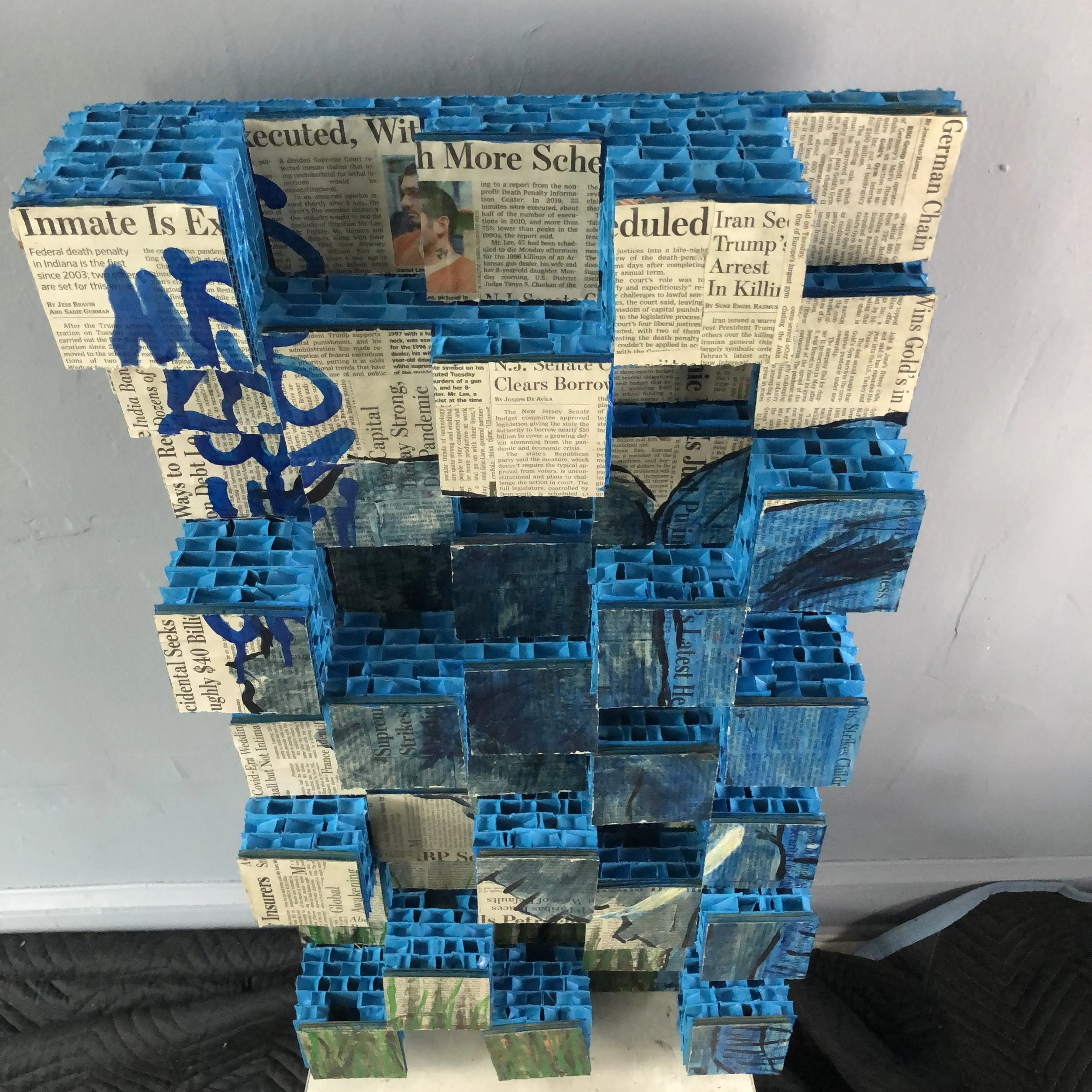 A mixed-media 3D collage art piece composed of newspaper-covered cardboard squares arranged in an abstract design, with a blue colored elephant in tall grass. Handwritten logo for "MISAKOSUZUKI" on the upper left in blue ink.