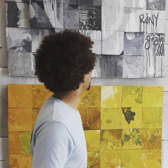 Person with curly hair viewed from the side, wearing a white shirt, standing in front of a wall with various colored tiles and artwork. MISAKOSUZUKI, MISAKO SUZUKI