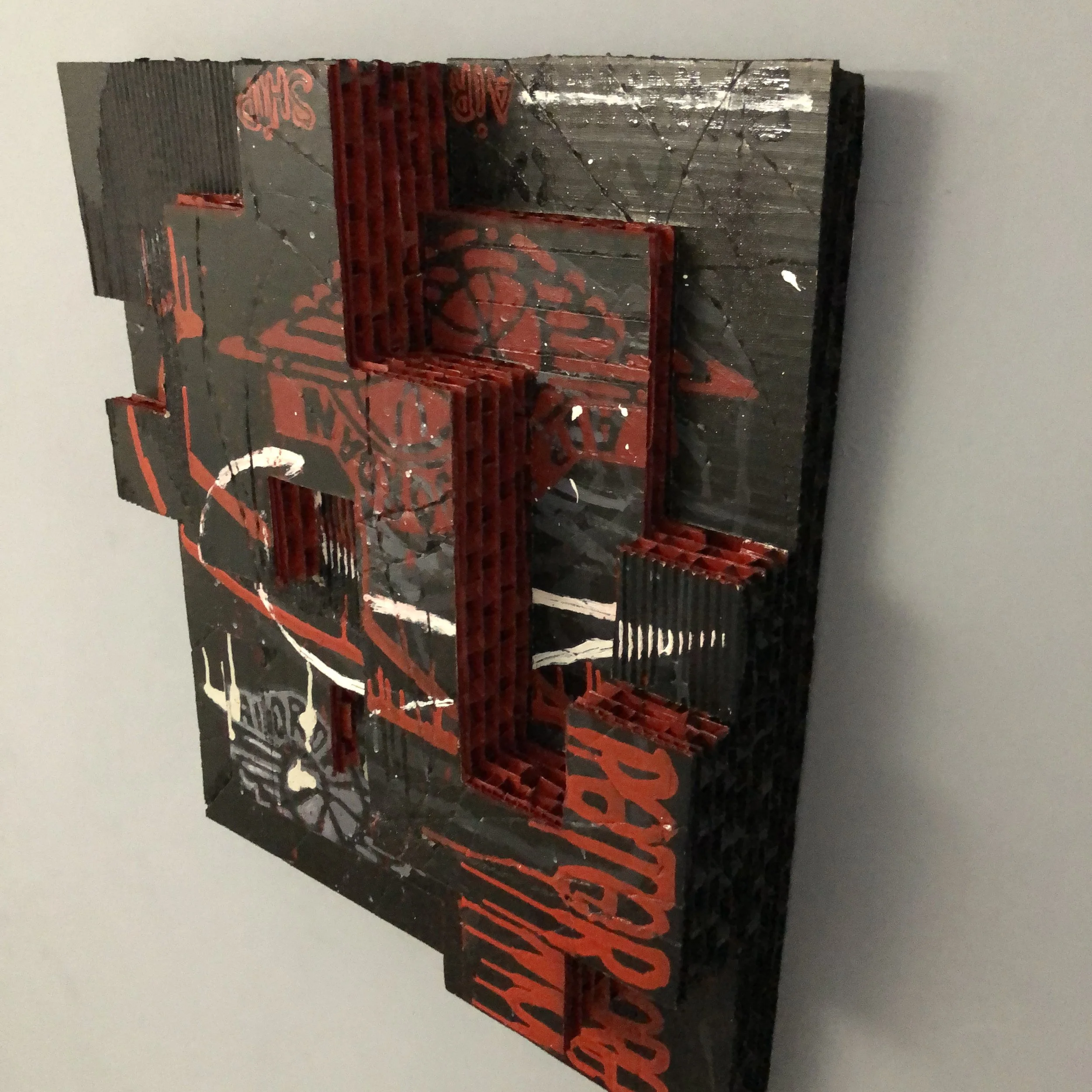A mixed-media art piece using cardboard divided into Tetris like shapes at differing heights to give the illusion of depth. Black background with red and white markingS and "NIKE" logo's. The name of a Nike designer "PETER" lower right in red ink.