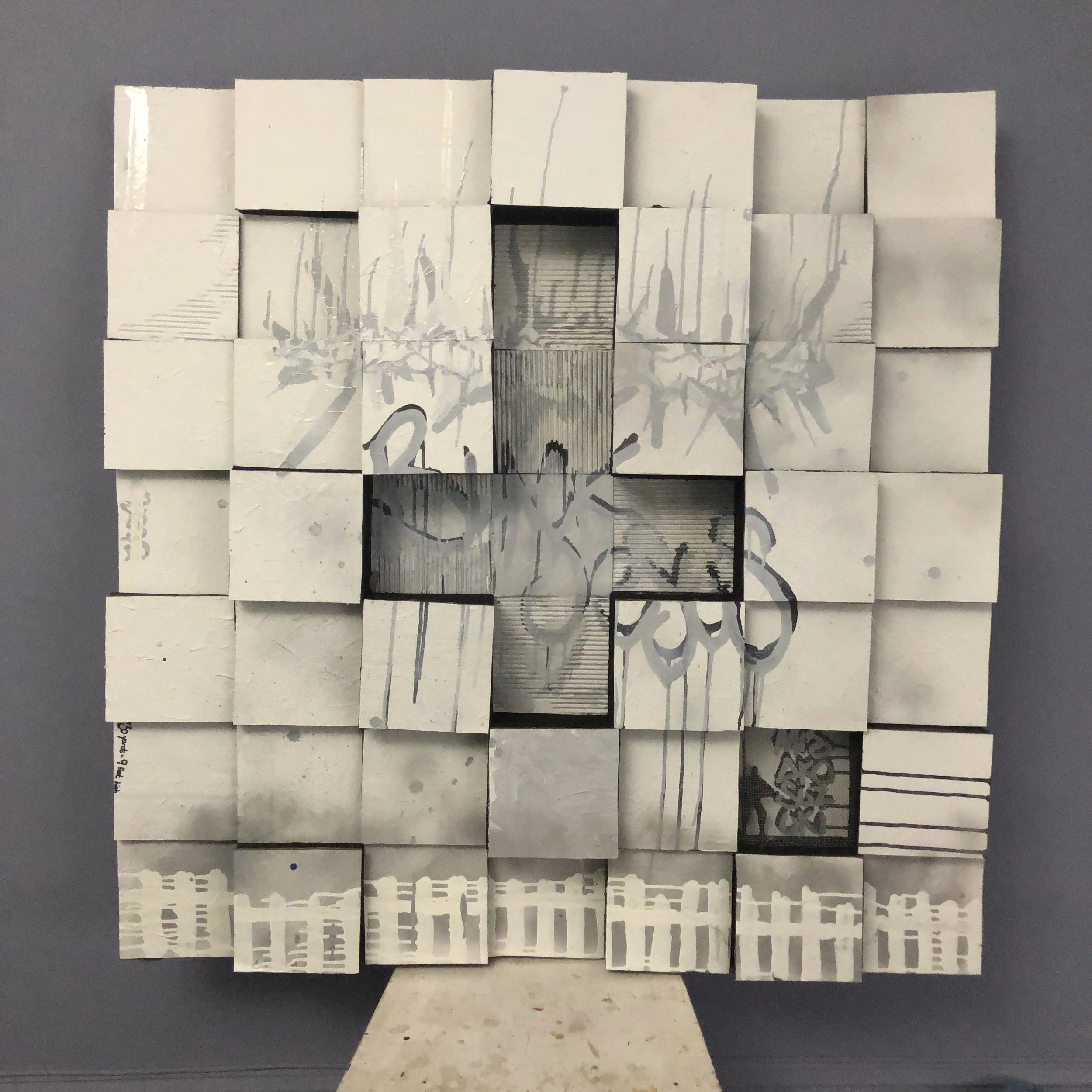 A large grid using cardboard painted white squares, an upside down crucifix design is recessed in the center and the word "BLACK JESUS" in the center.The bottom has a row of white fences in ink and there is also a crown of thorns in silver ink.