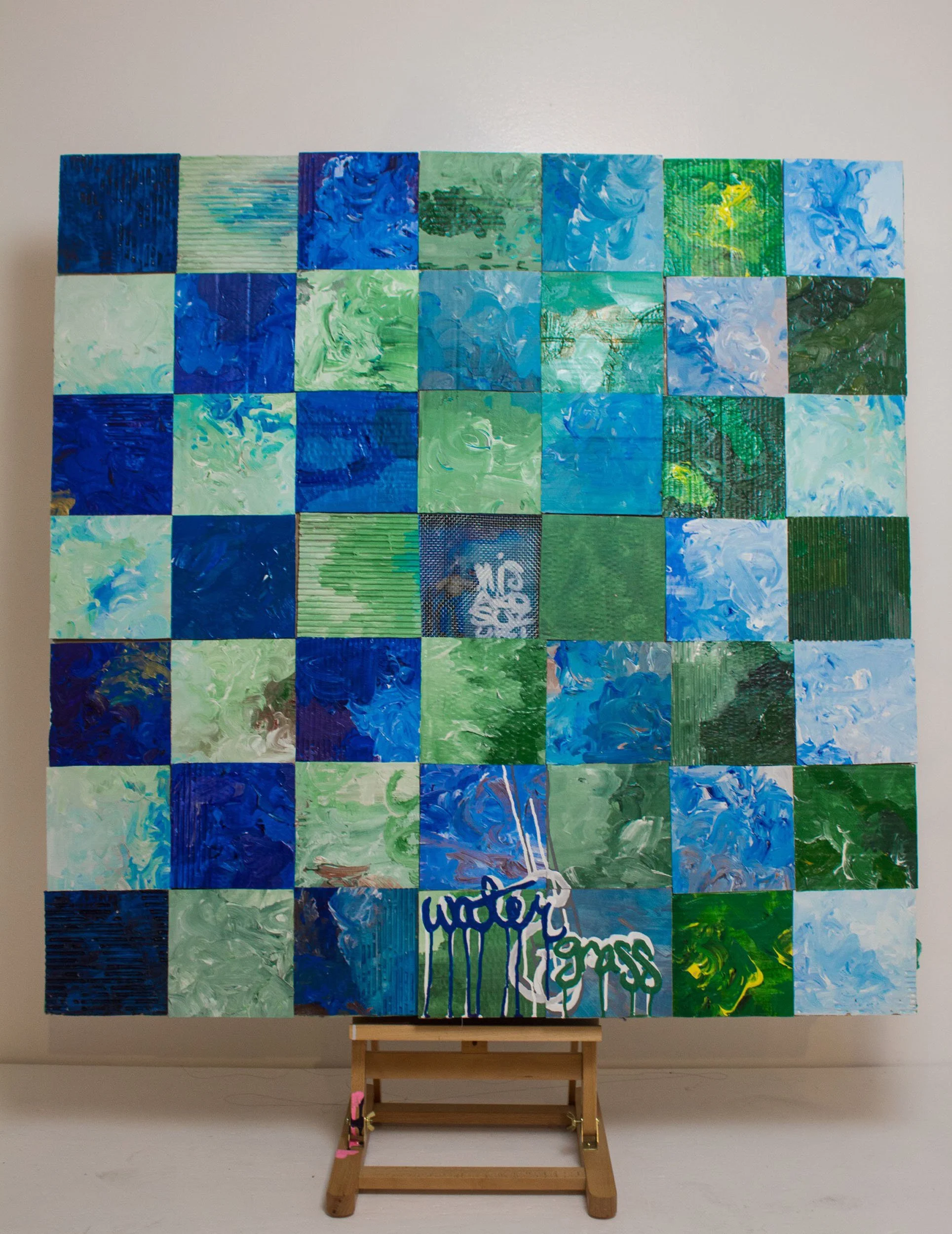 A mixed-media, grid-like art piece composed of cardboard squares arranged in a checkerboard design, with alternating blue and green squares.