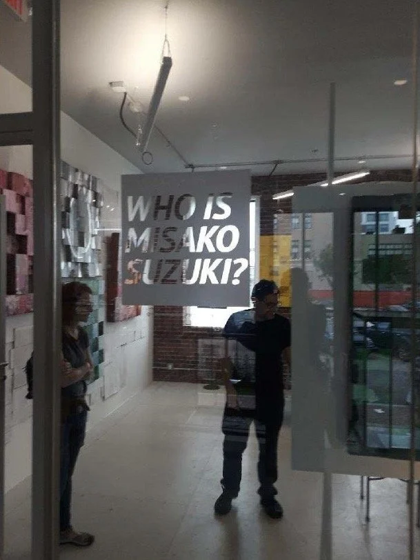 Interior view of a modern building lobby with a sign that reads 'Who is Misako Suzuki?' visible through glass. Art exhibition of MISAKO SUZUKI, or MISAKOSUZUKI; Boston, MA exhibition