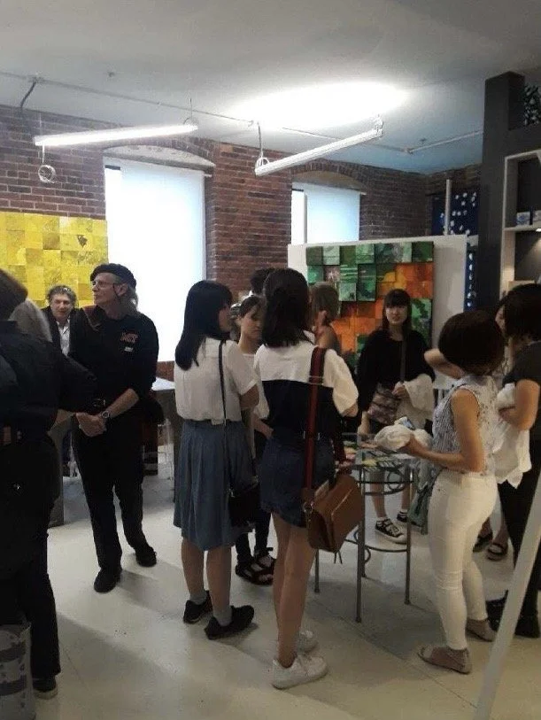 Group of people gathered around a table displaying items in a room with brick walls, colorful artwork, and large windows. MISAKO SUZUKI, MISAKOSUZUKI
