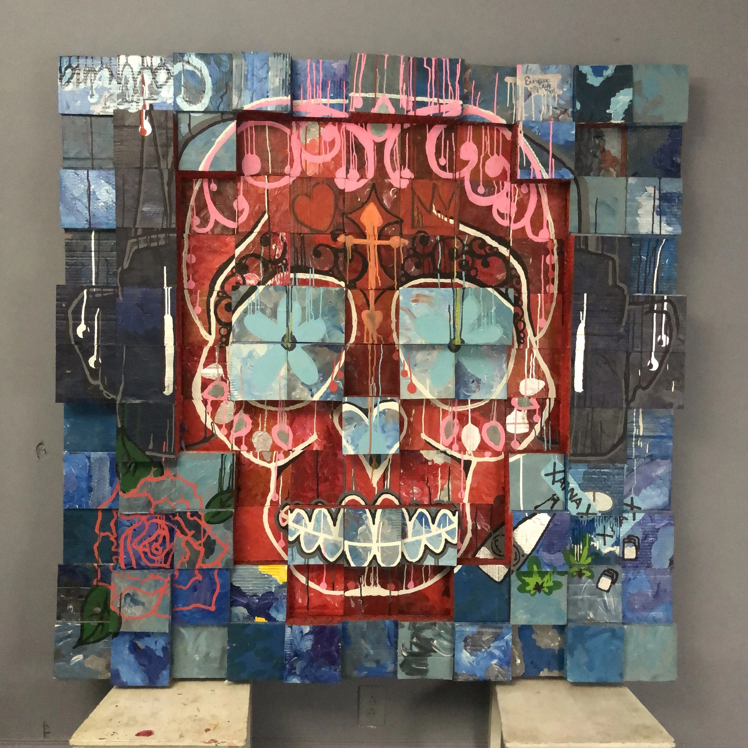 A mixed-media, grid-like art piece composed of cardboard squares arranged in an abstract design, with a red colored skull in the center surrounded by blue. With blue pills labeled "XANAX" painted on the lower right section and a flower on the left..