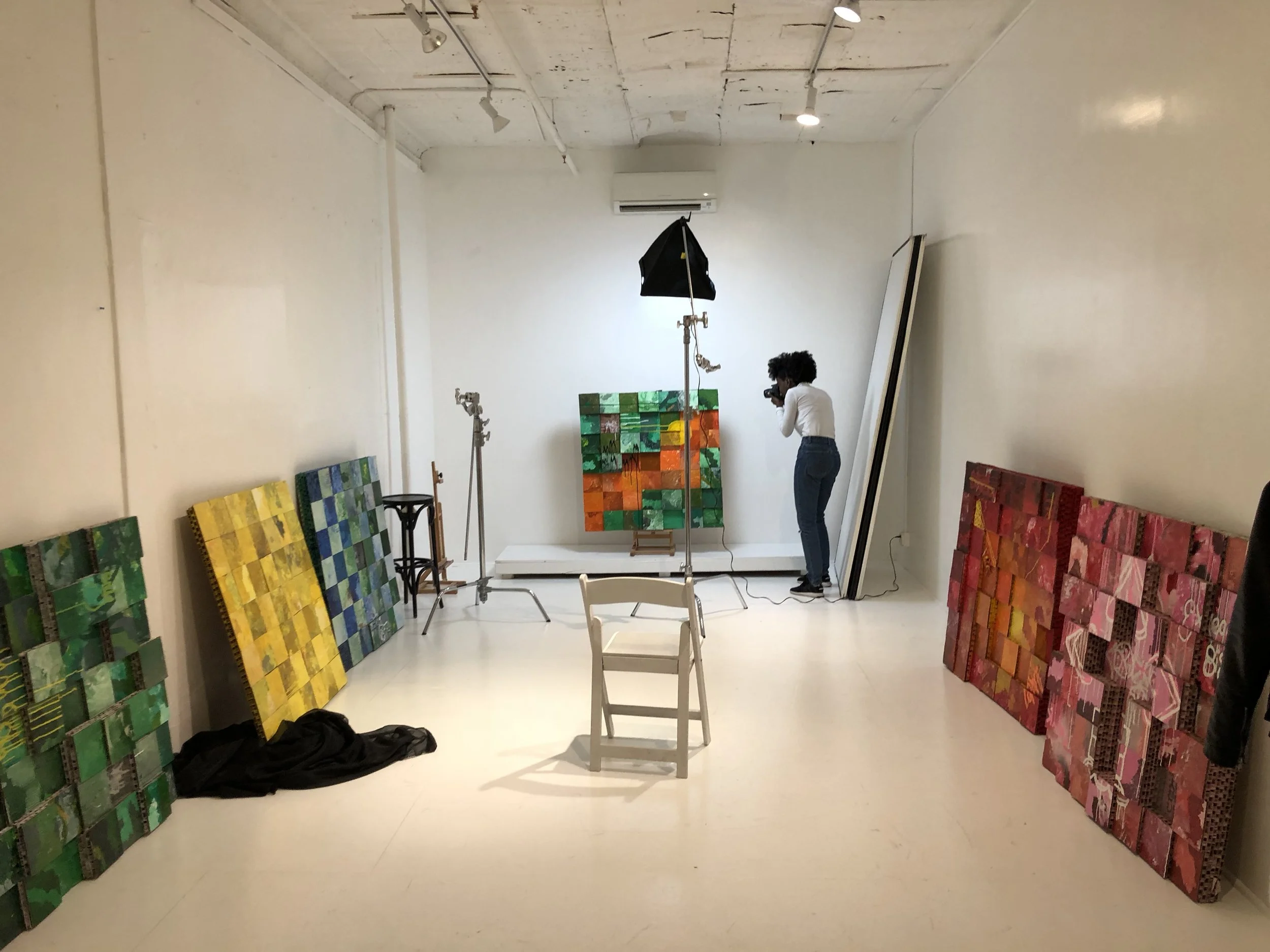 An artist's studio with colorful abstract paintings leaning against the walls, a photographer taking pictures of a green and orange artwork on a white platform, and photography equipment including lighting and tripods.