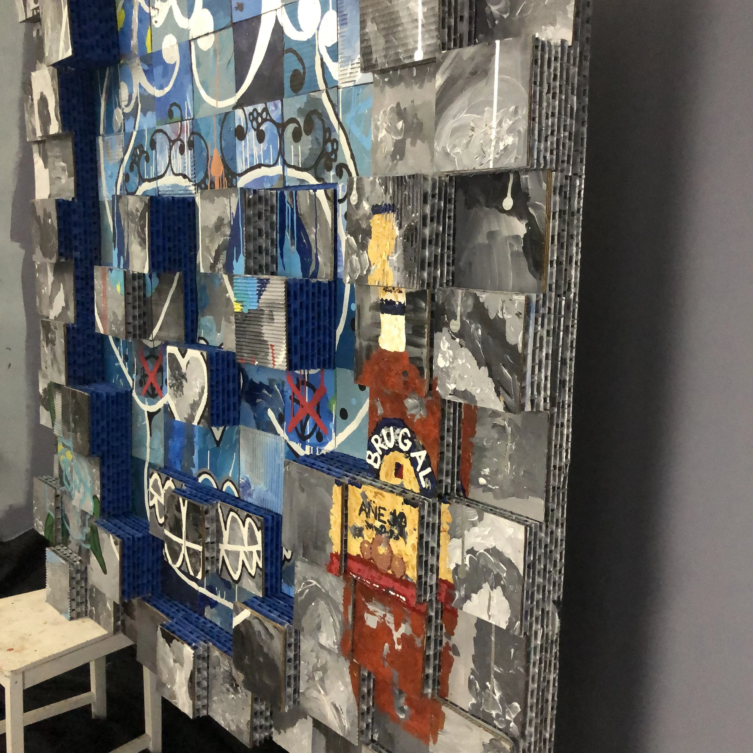A mixed-media, grid-like art piece composed of cardboard squares arranged in an abstract design, with a blue colored skull in the center surrounded by gray. With a bottle of "BRUGAL" painted on the lower right section and a flower on the left..