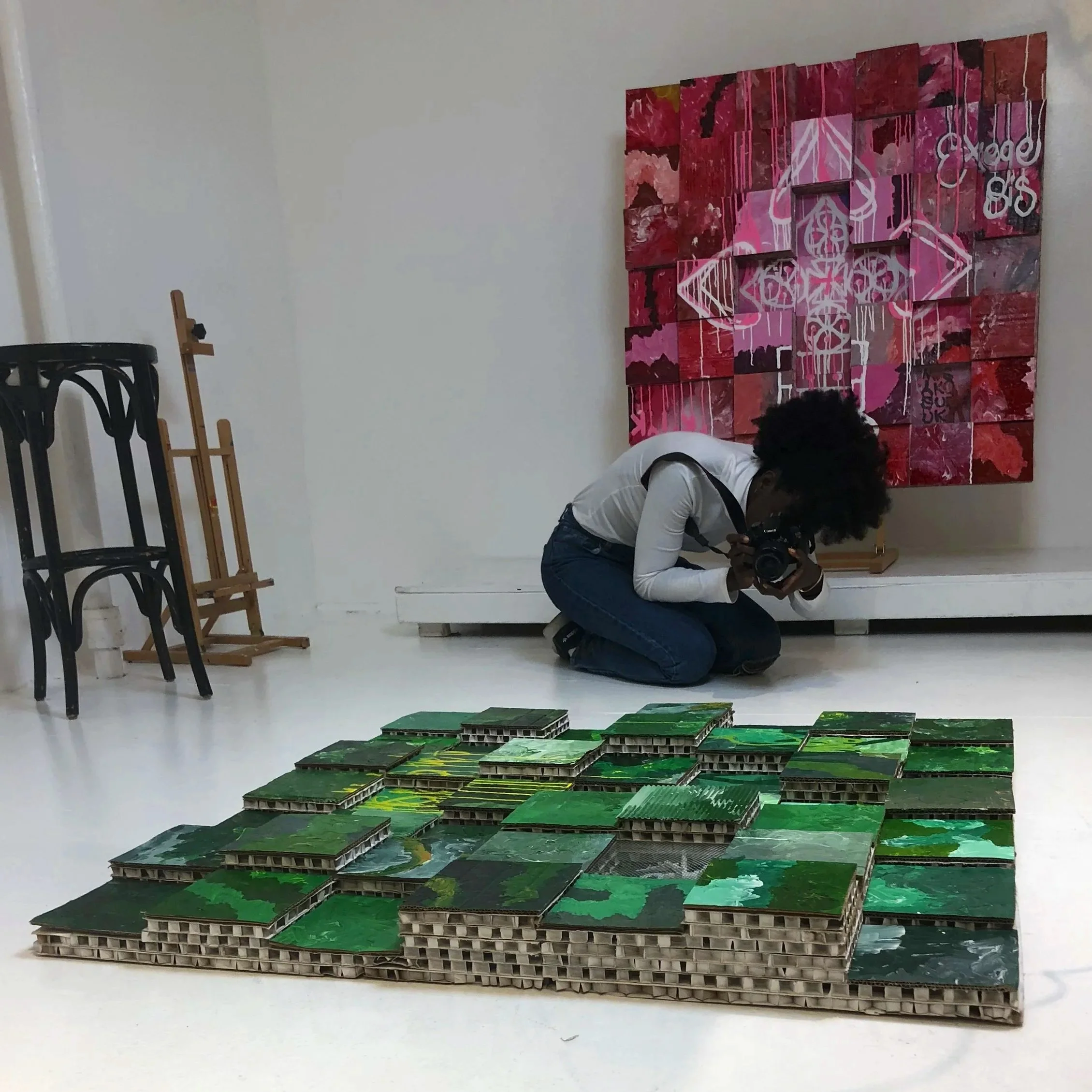 Artist kneeling on the floor taking a photograph of a mixed media artwork. The artwork consists of small three-dimensional pieces arranged in a grid, painted with various shades of green. In the background, there is another larger artwork on the wall, with pink, red, and white colors, and abstract patterns.