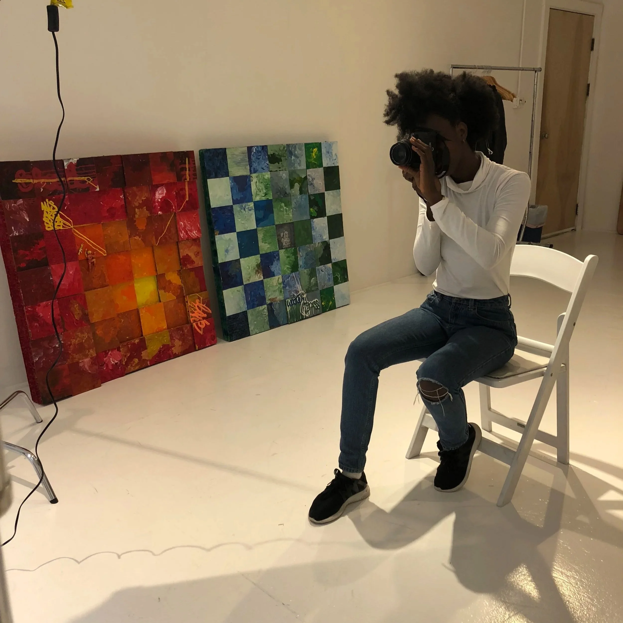 A person sitting on a white chair taking a photo in front of a mirror. There are two large abstract paintings leaning against the wall, one with red and yellow tones, and the other with green and blue tones. A black cord hangs from the ceiling.