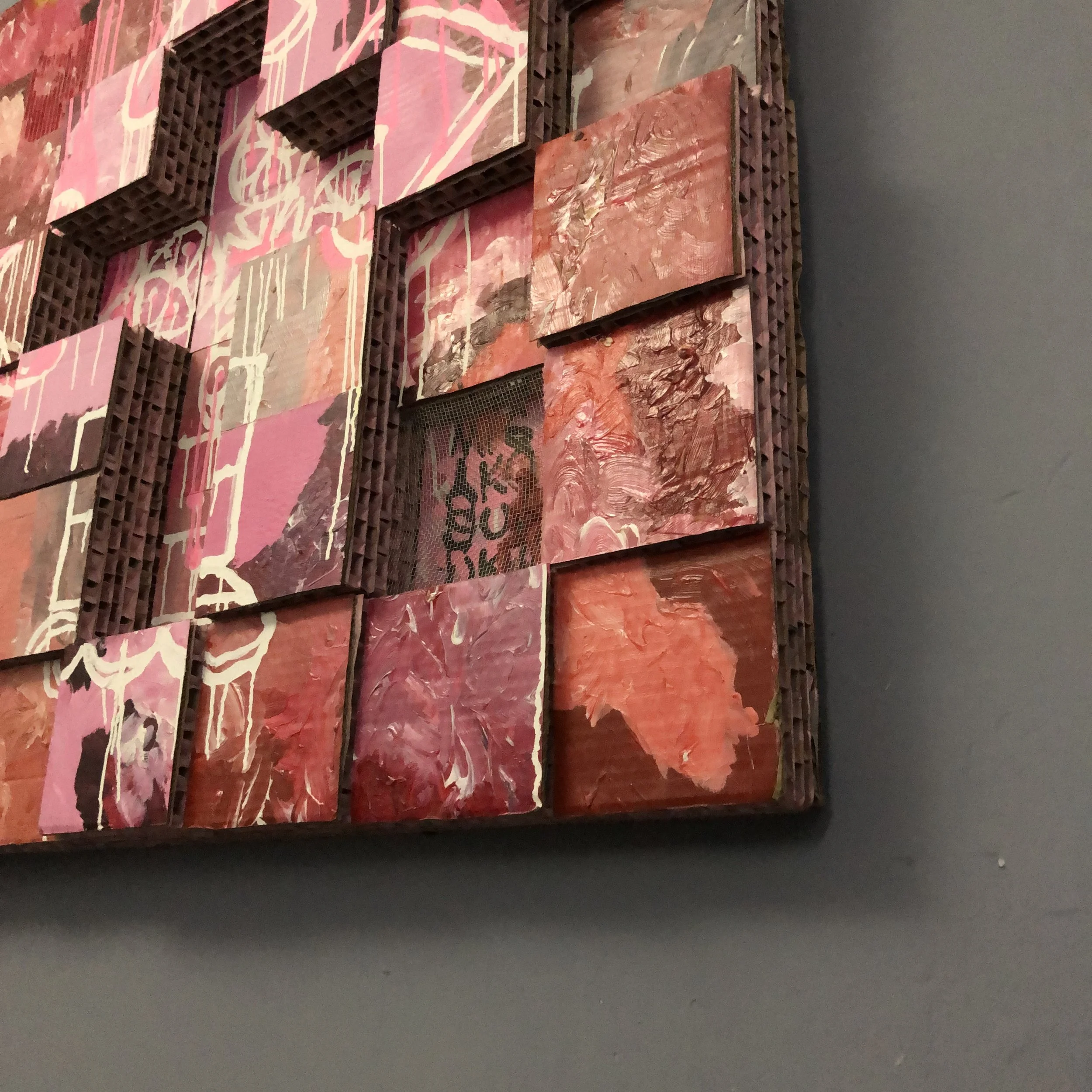 Close-up of abstract mixed media artwork with pink, red, and purple colors, created with paint and textured elements mounted on a dark wall.