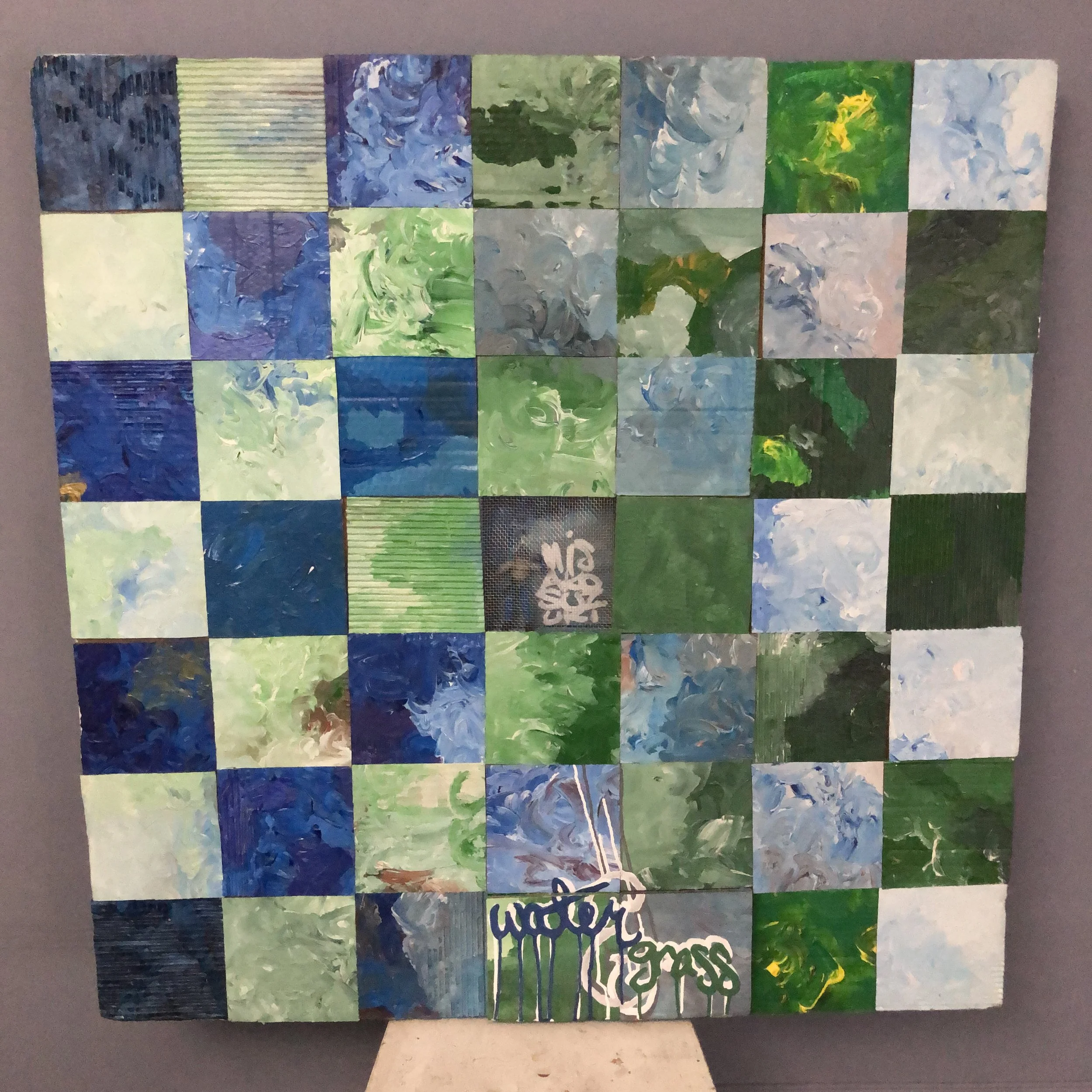 A mixed-media, grid-like art piece composed of cardboard squares arranged in a checkerboard design, with alternating blue and green squares.