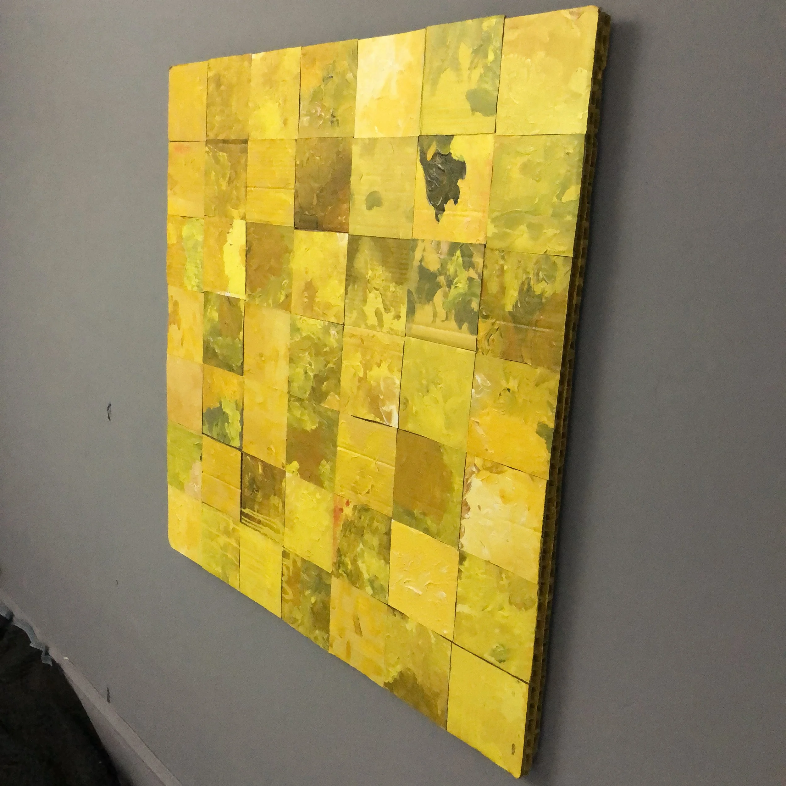 A square mosaic artwork composed of small yellow and green painted blocks arranged on a gray wall.