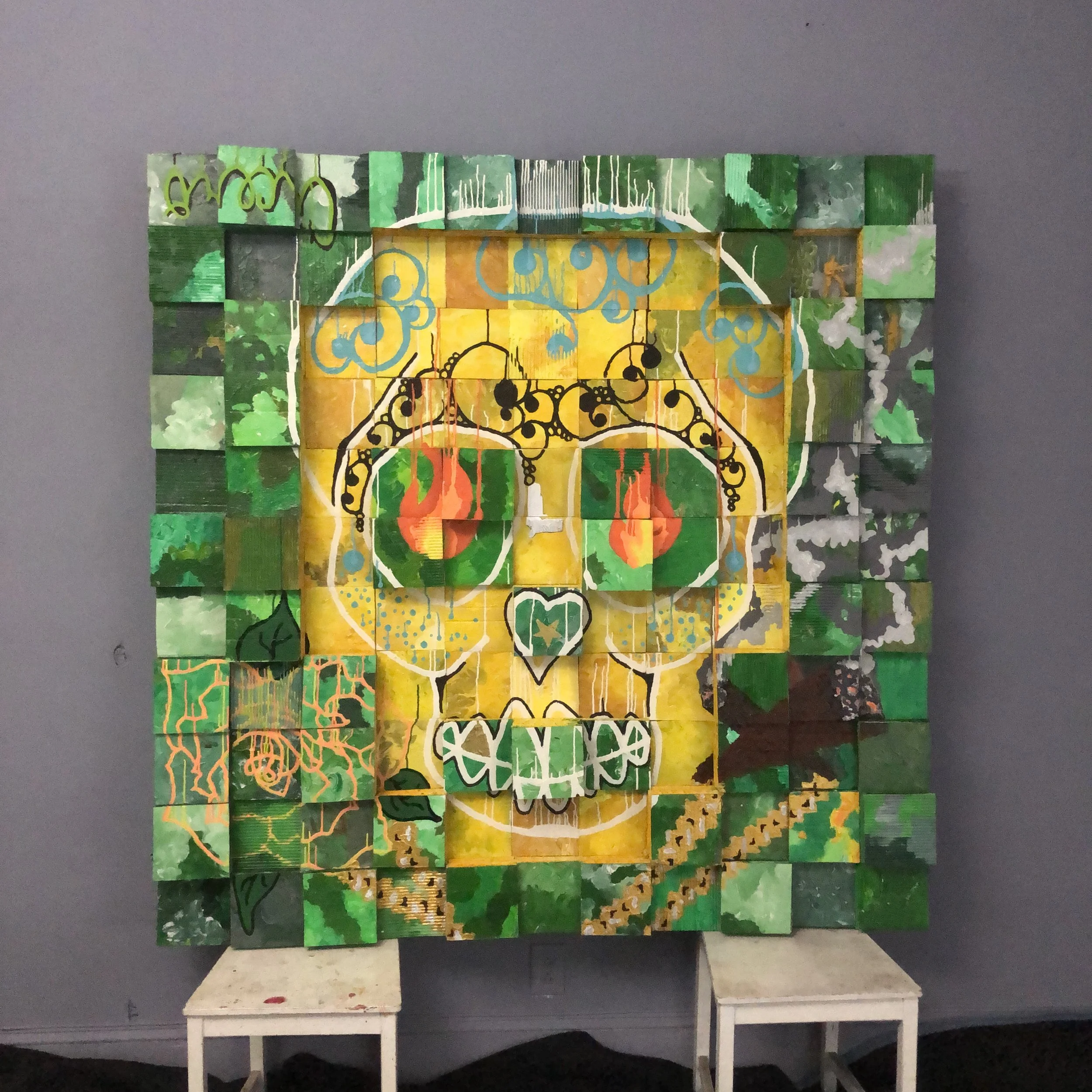 A mixed-media, grid-like art piece composed of cardboard squares arranged in an abstract design, with a yellow colored skull in the center surrounded by green. With brown cigars burning painted on the lower right section and a flower on the left..