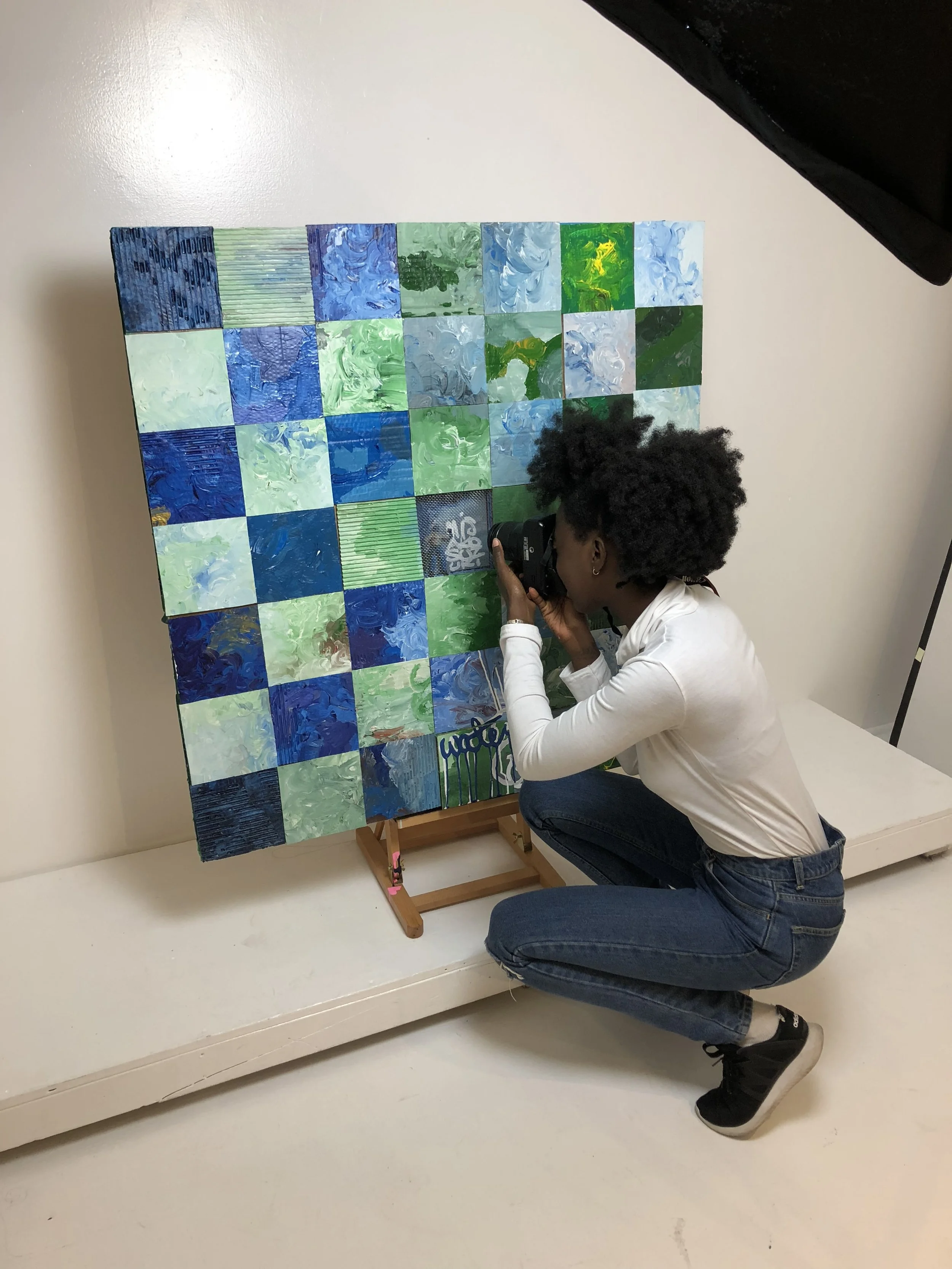 A woman in a white long-sleeve top and jeans is crouching and taking a photo of a colorful abstract floral artwork composed of several squares with blue, green, and light colors on an easel.