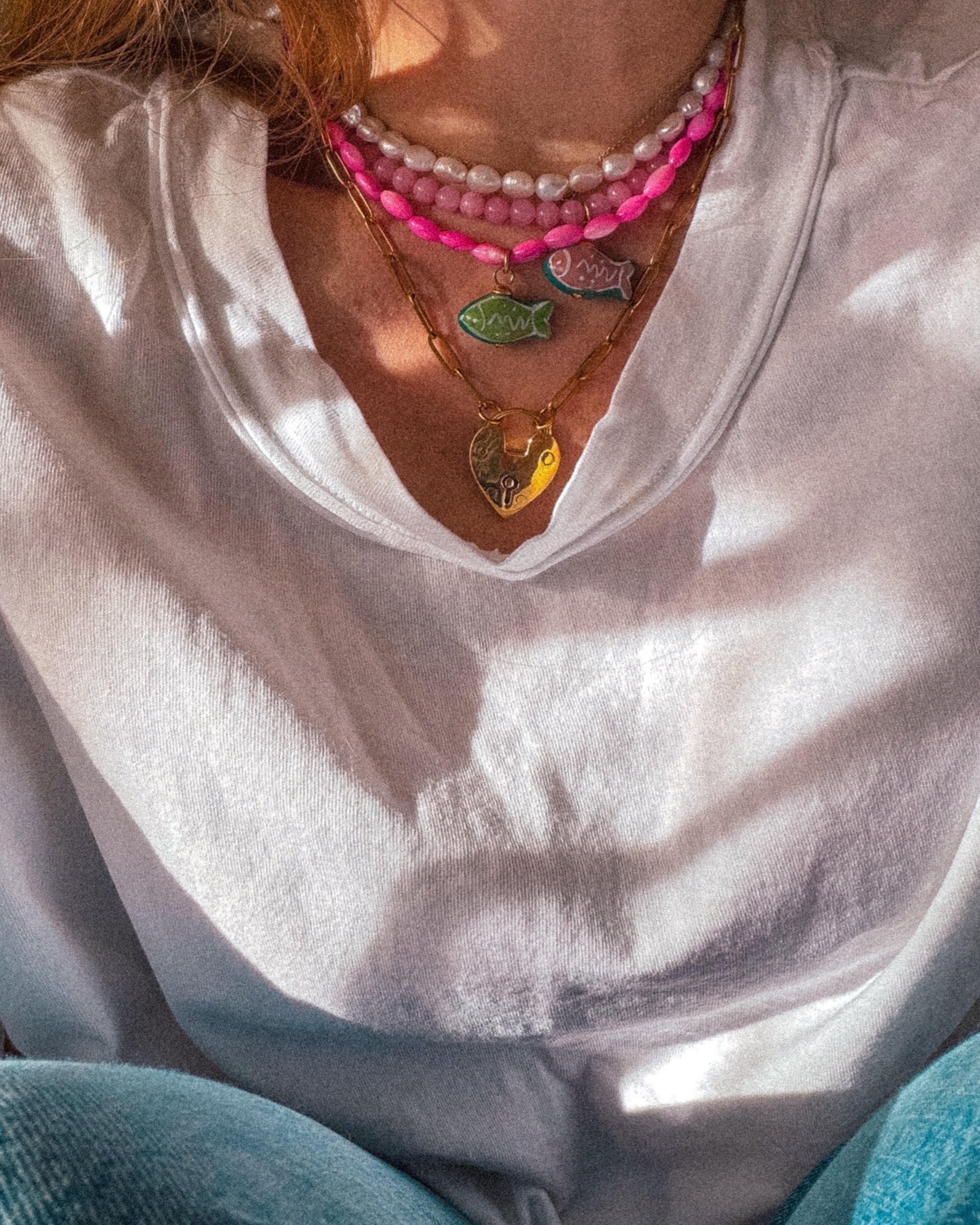 Close-up of a person's neck and chest wearing layered necklaces with fish-shaped pendants, a cream-colored shirt, and blue jeans.