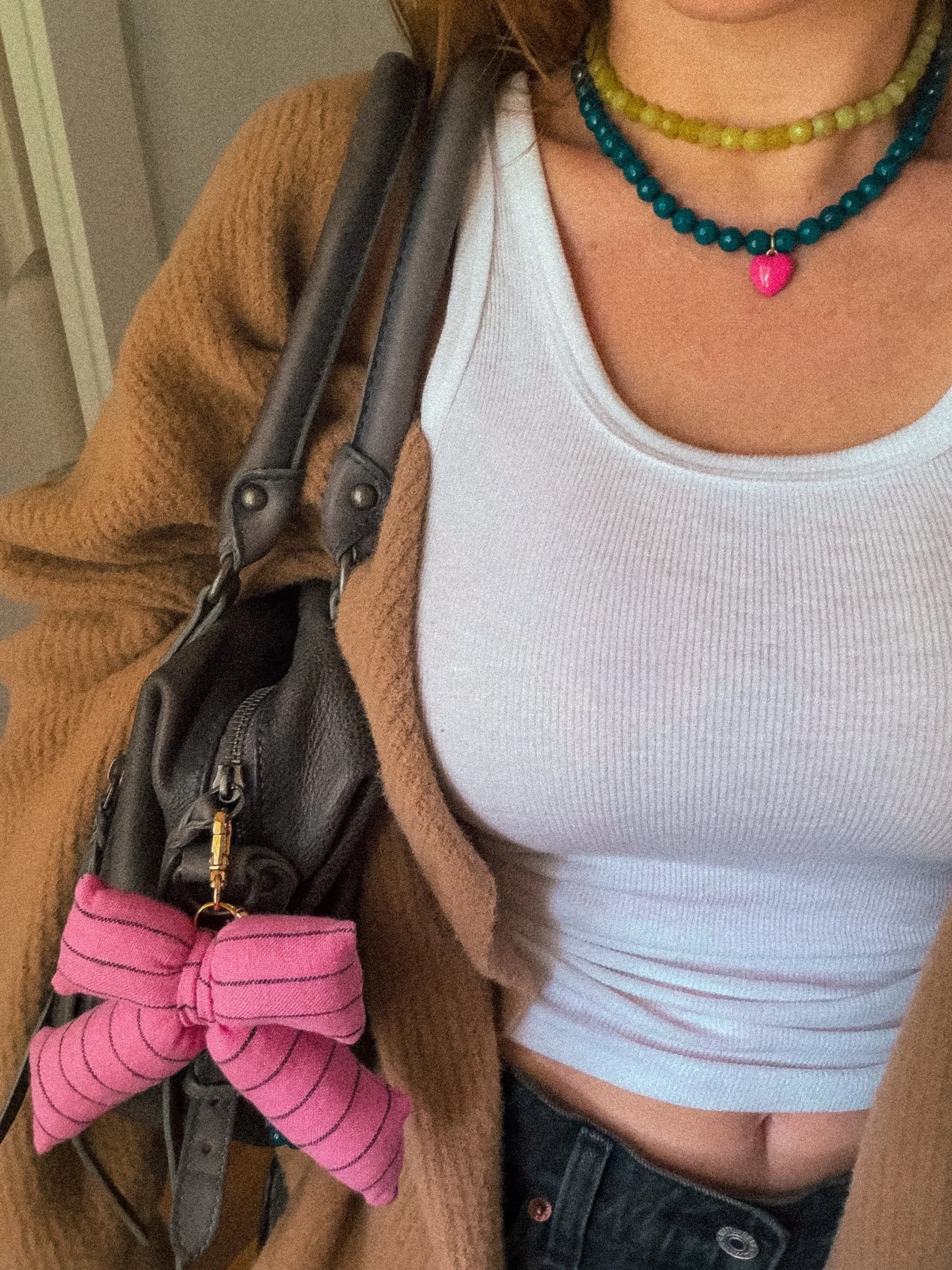 Puffy pink bow bag charm with dark stripes clipped to a gray leather bag, styled with a white tank top and teal beaded necklace