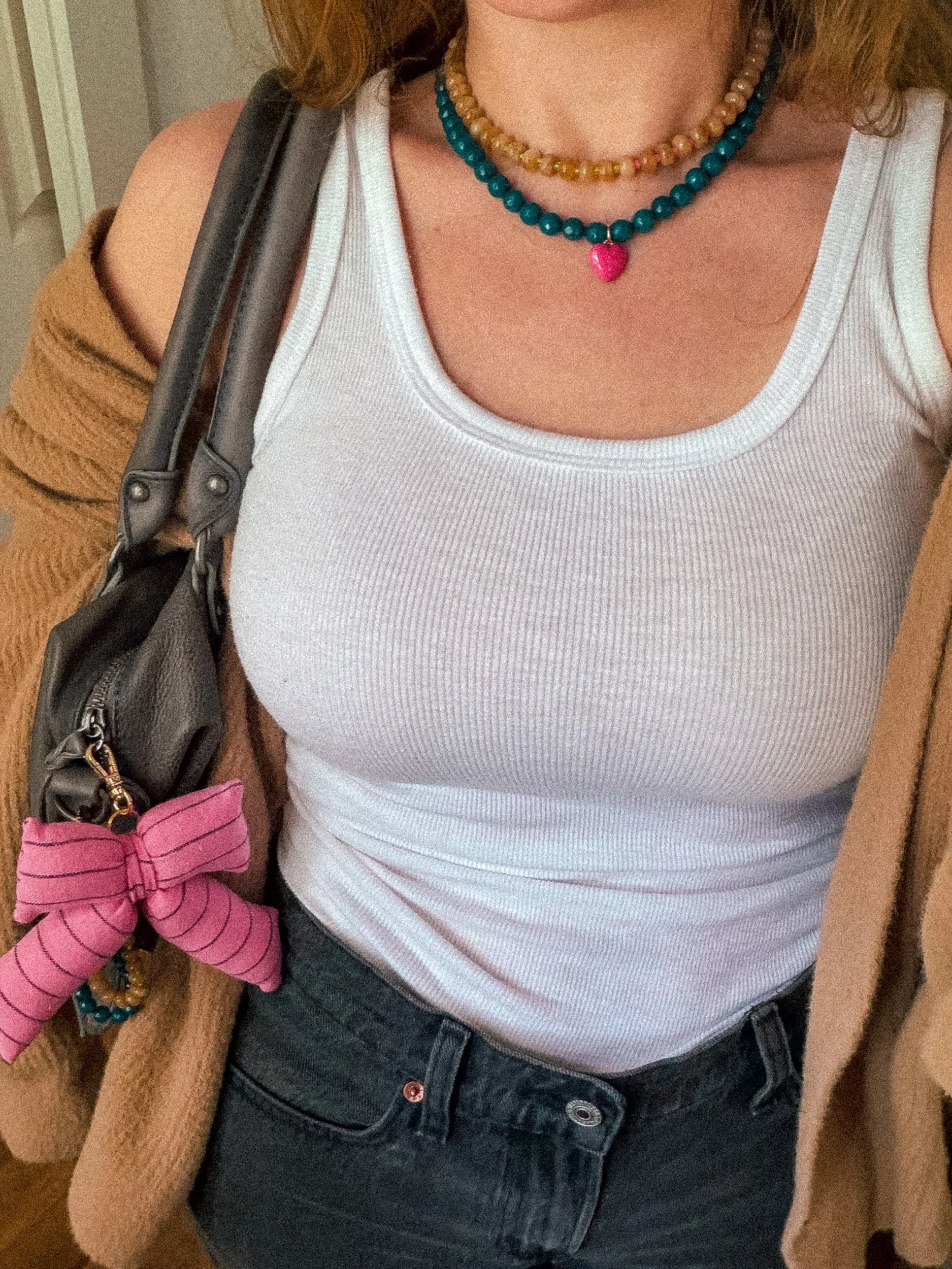 Handmade striped bow bag charm in pink hanging from a gray leather handbag, worn with a white tank top and brown cardigan