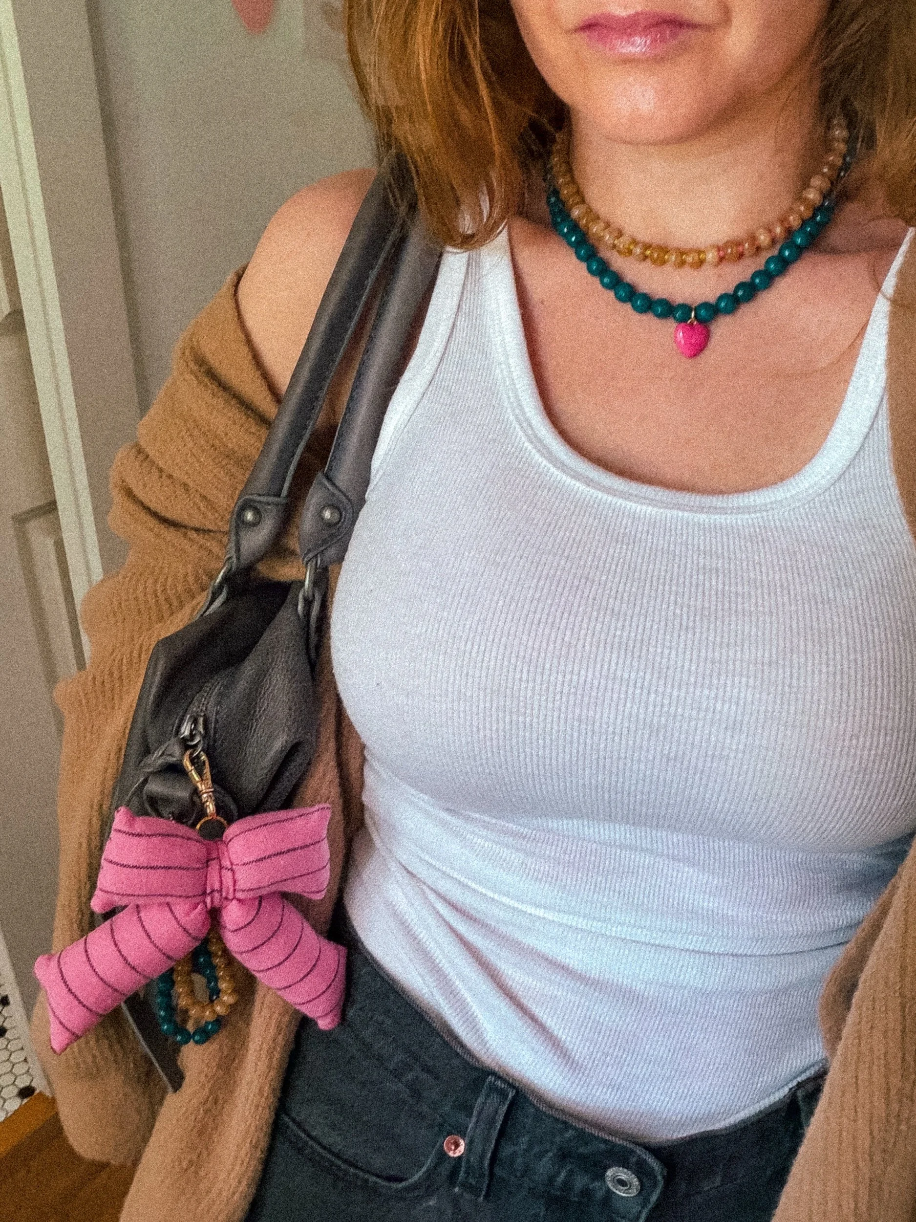 Amber and tan beaded necklace layered with a teal heart charm necklace, worn with a white tank top, brown cardigan, and pink striped bow bag charm