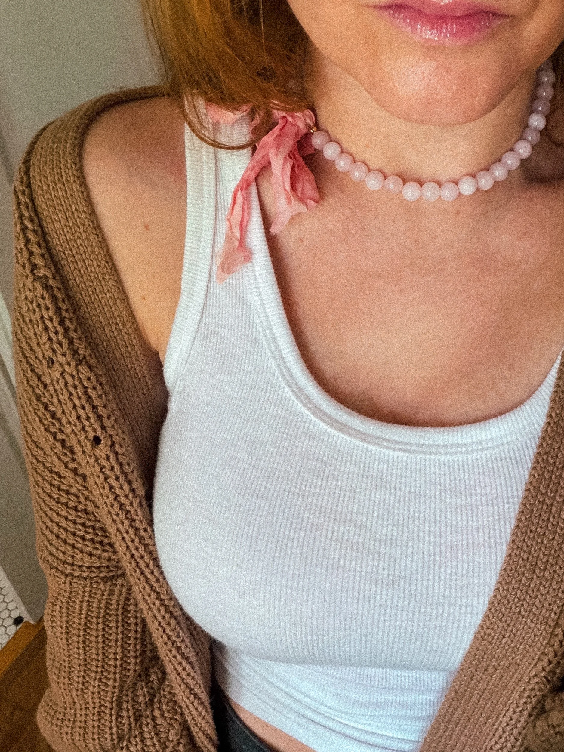 Pale pink beaded necklace with pink ribbon bow worn with a white ribbed tank top and brown knit cardigan