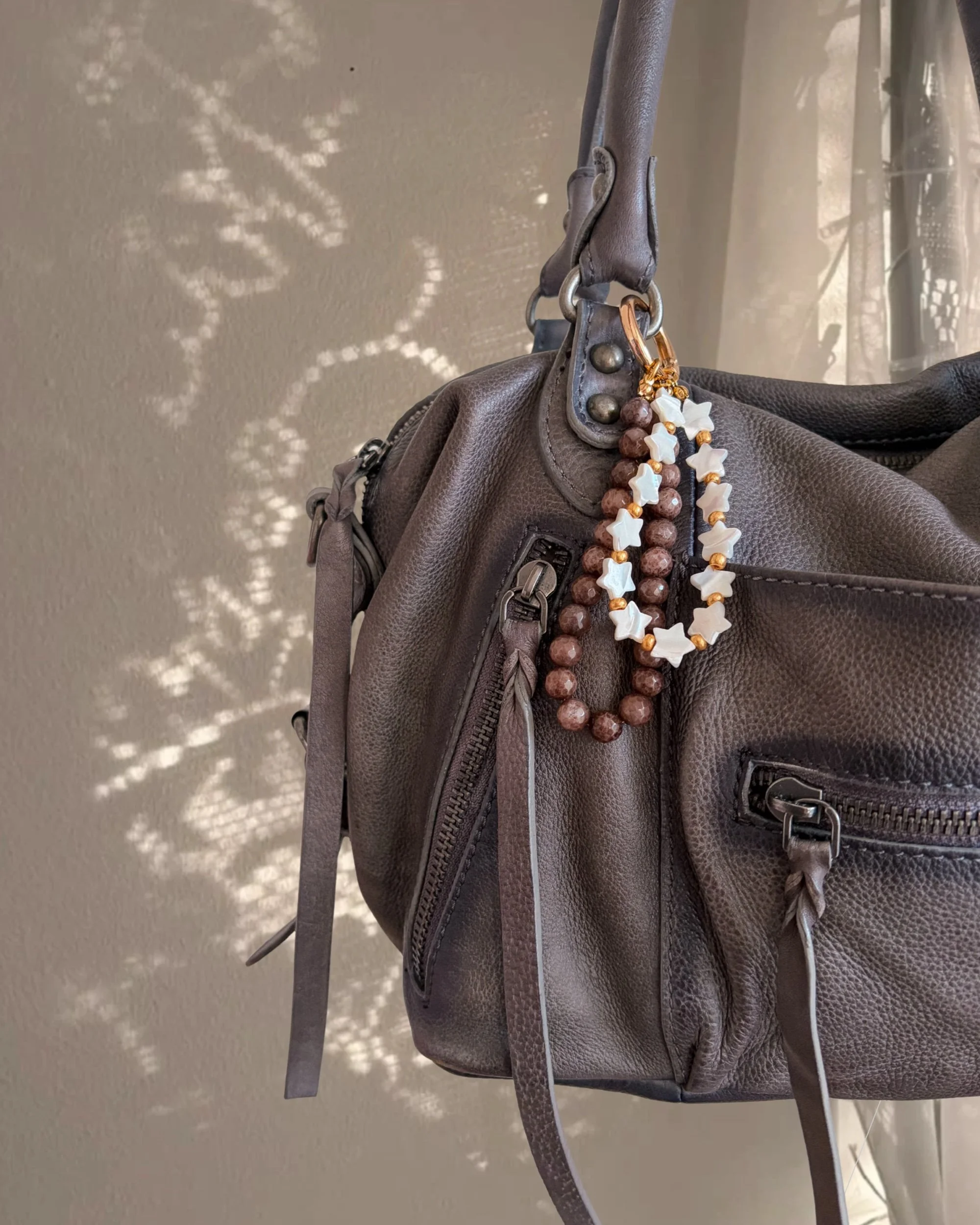 Brown and white star bead bag charm hanging from a gray leather handbag, with sparkly star-shaped light behind