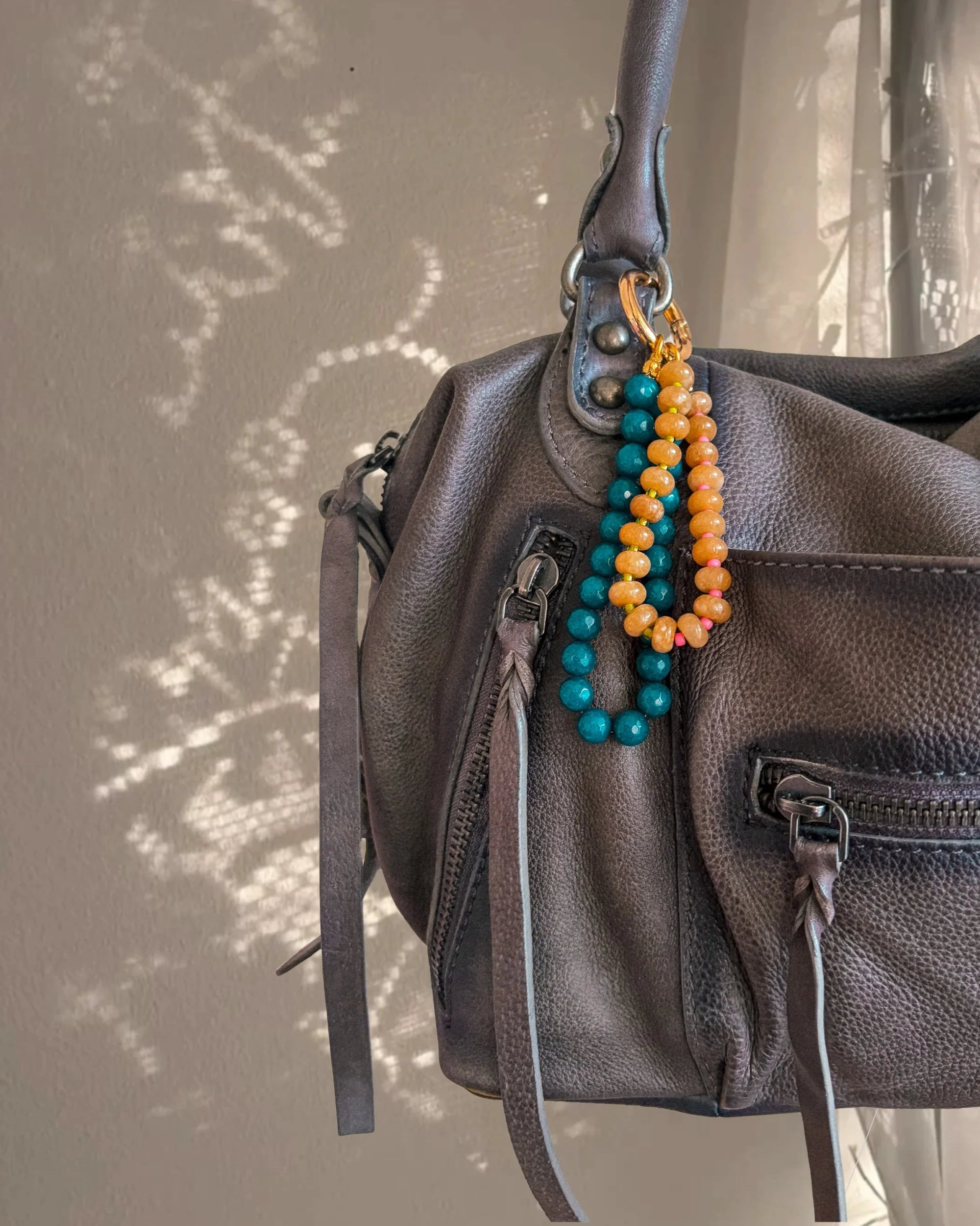 Teal and amber beaded bag charm hanging from a gray leather handbag, with sparkly star-shaped light behind
