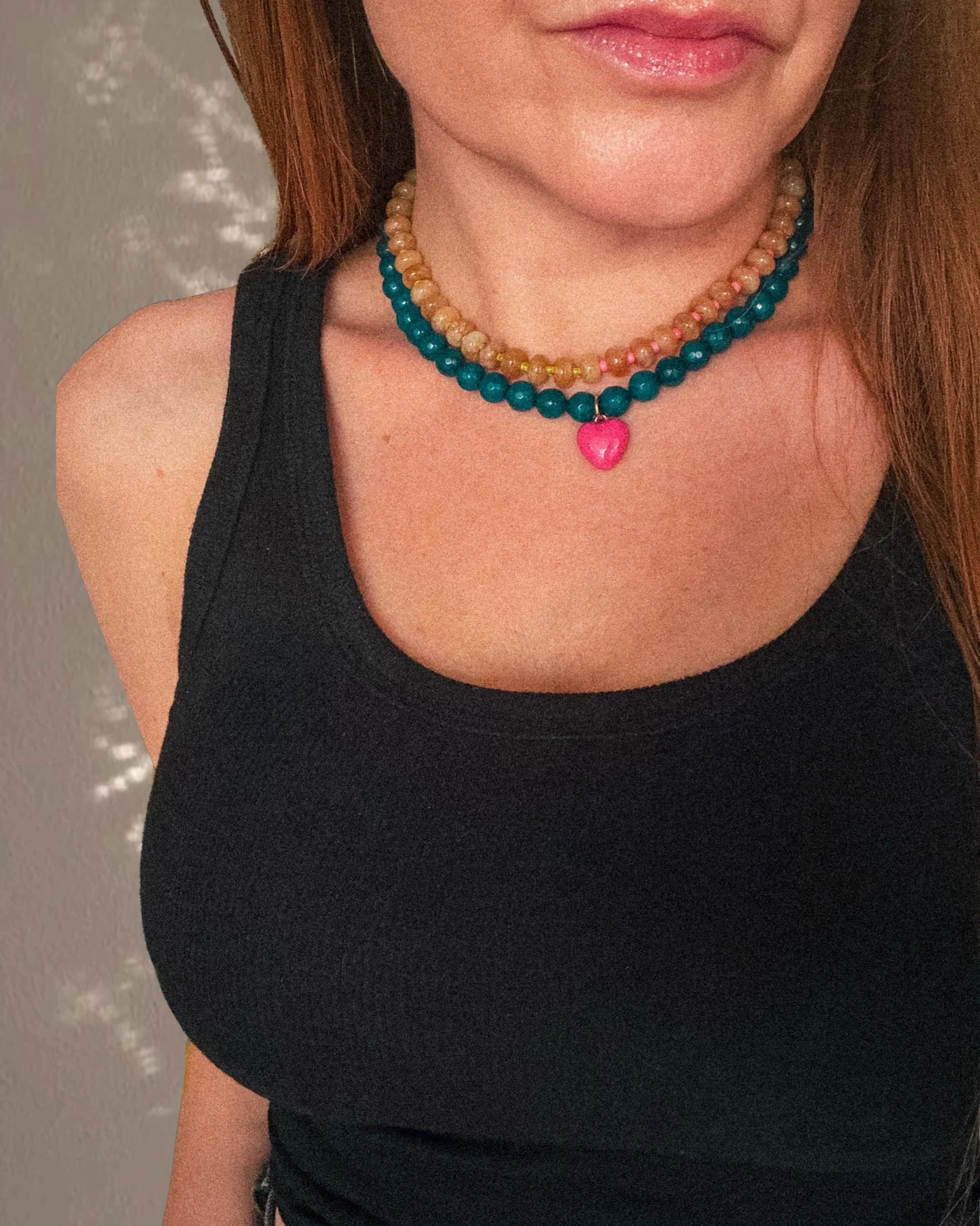 The image shows a woman’s neck and lower face wearing three layered necklaces, the top one has light-colored beads with a pink heart pendant. She is wearing a black sleeveless top and has long reddish-brown hair.
