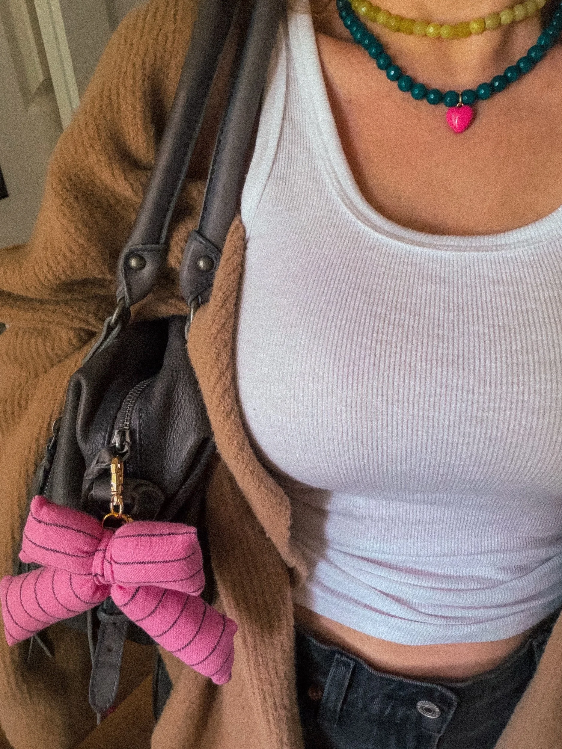 Close-up of a person's chest and shoulder showing layered necklaces, a white ribbed tank top, a brown and black jacket, and part of a black purse with a pink bow detail, all on a beige textured sofa.