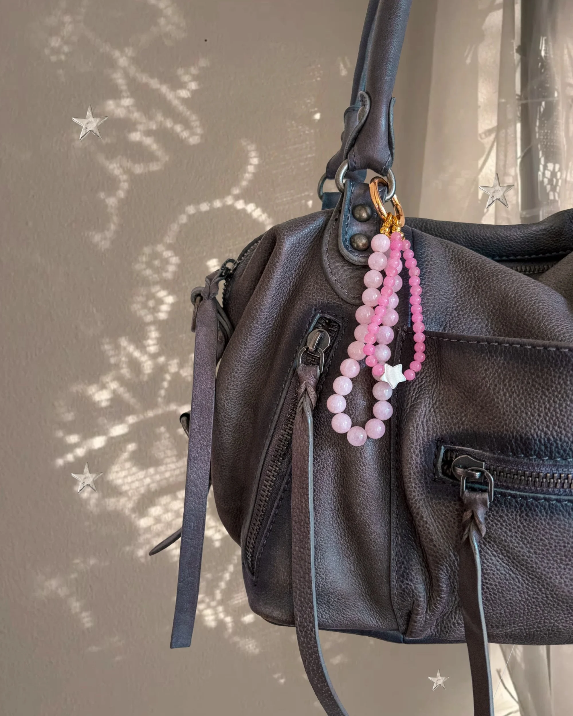 Pink beaded bag charm with white star bead hanging from a gray leather handbag, with sparkly star-shaped light behind