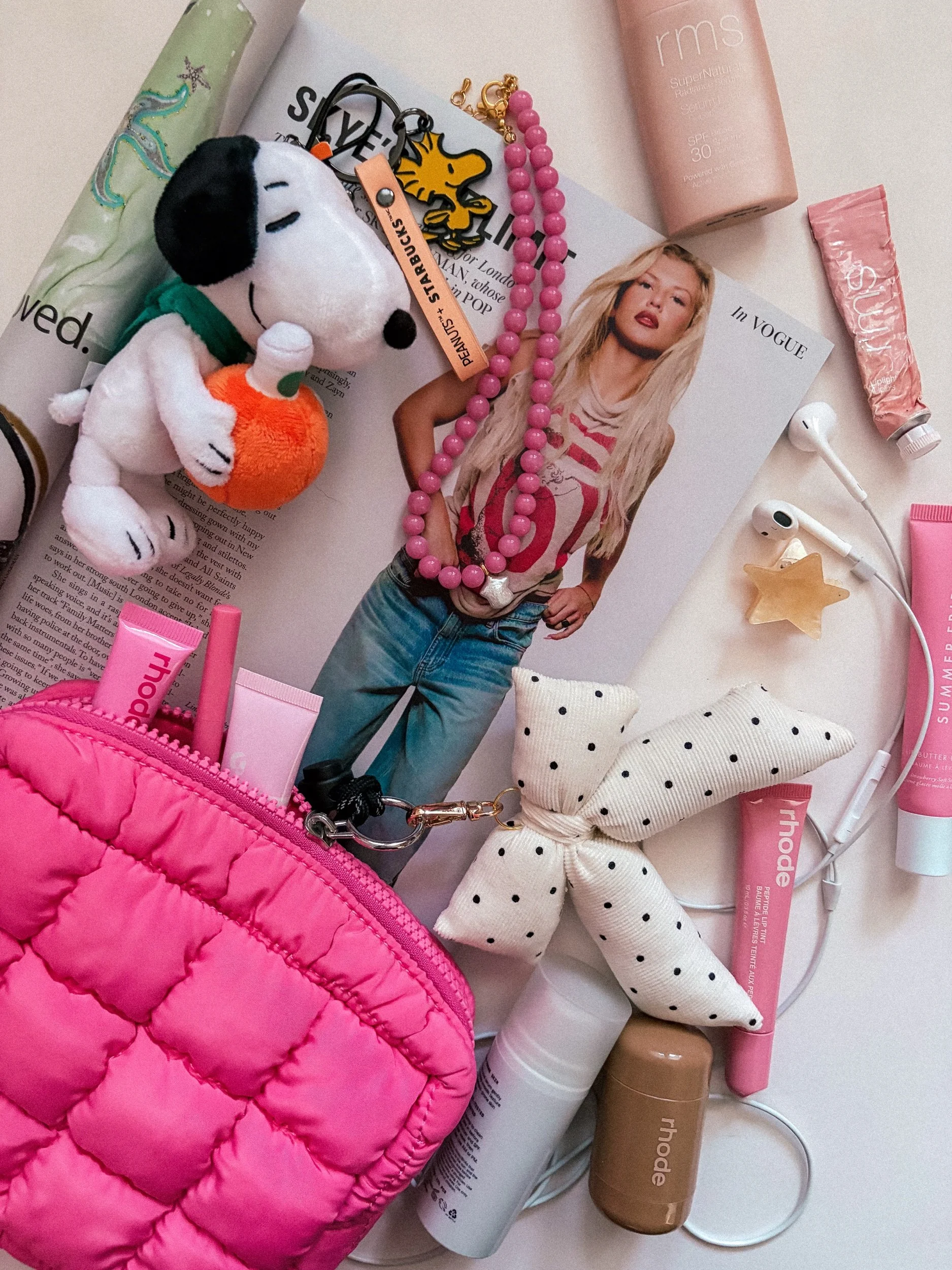 A collection of personal items including a plush Snoopy toy holding an orange plush, pink quilted bag, fashion magazine, pink beaded necklace, skincare products, and a pair of polka-dotted socks on a white surface.