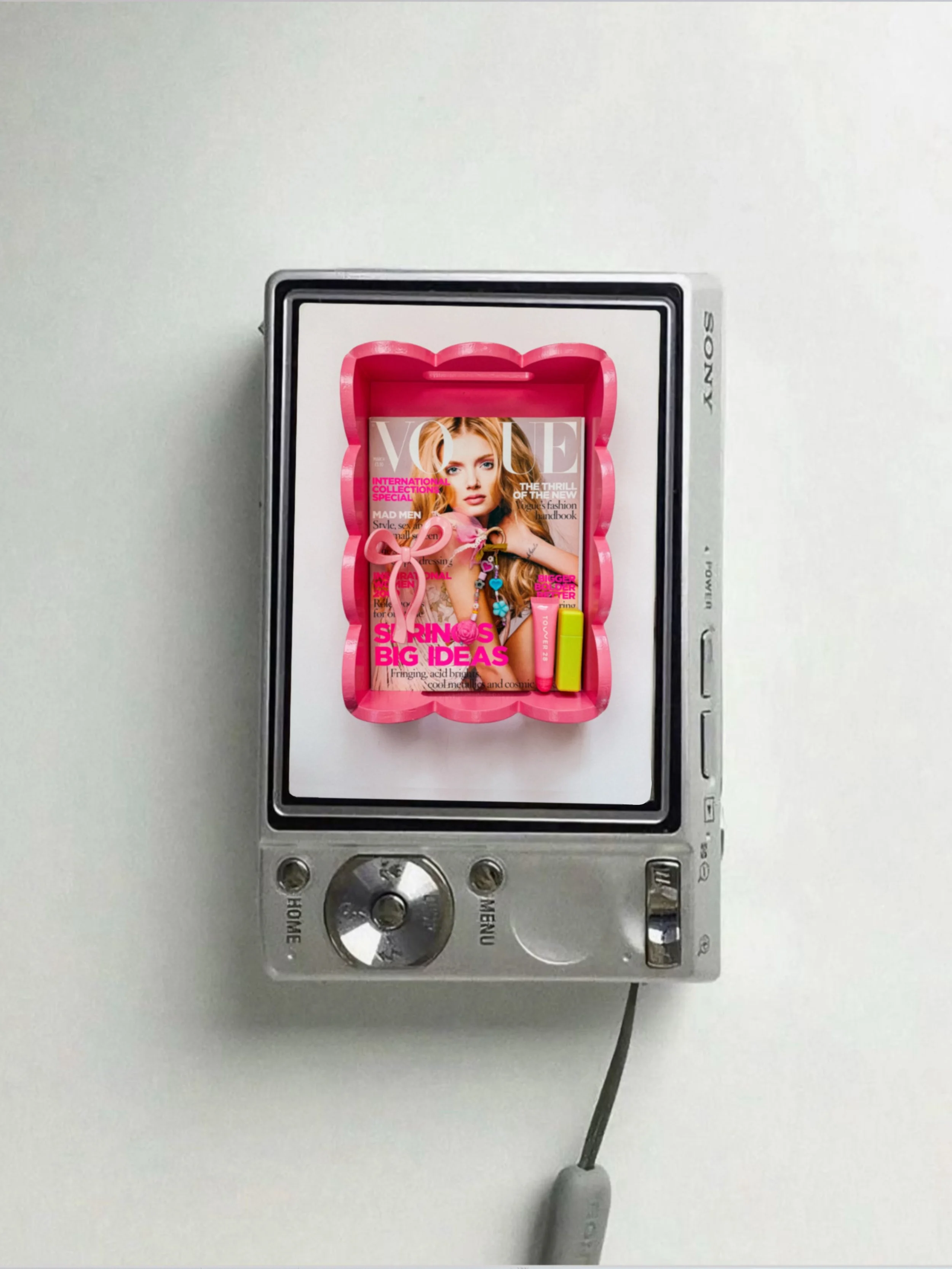 A metal control panel with a screen displaying a Vogue magazine cover featuring a red-haired woman, pink and yellow highlighters, and a pink bow-shaped frame, mounted on a white wall.