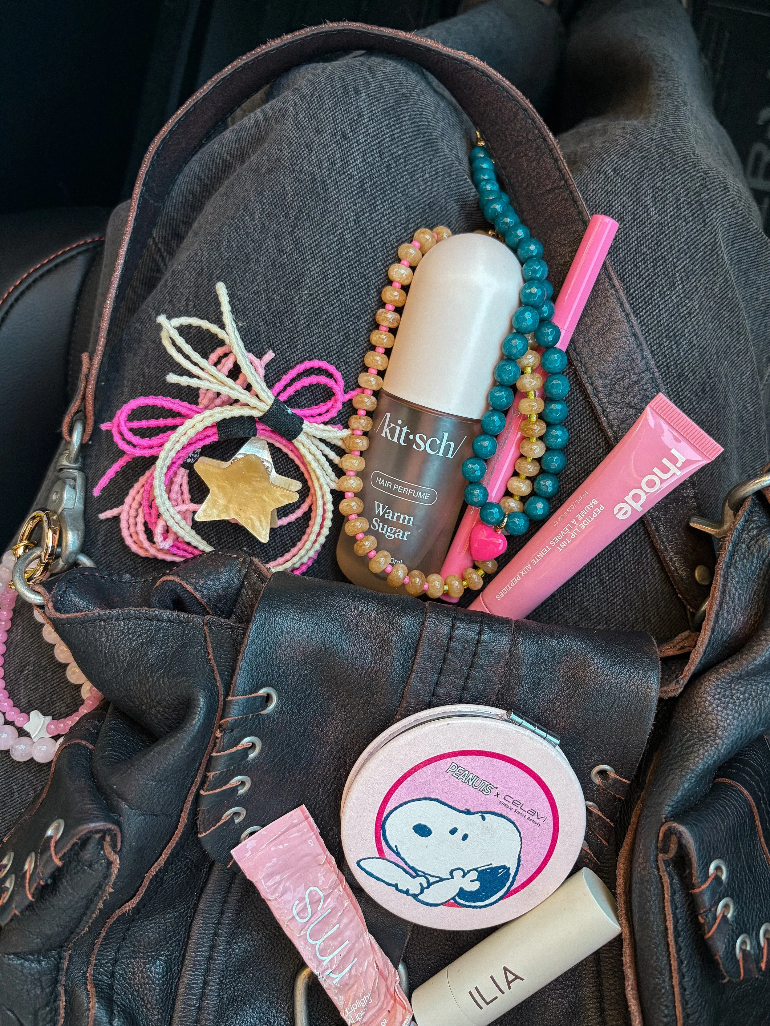 A black leather bag containing various items including a beige tube of 'Ilia' skincare, a pink 'Andalou' tube, a white 'Peanuts + Clay' compact, colorful beaded necklaces, a star-shaped hair accessory, and a pink hair clip.