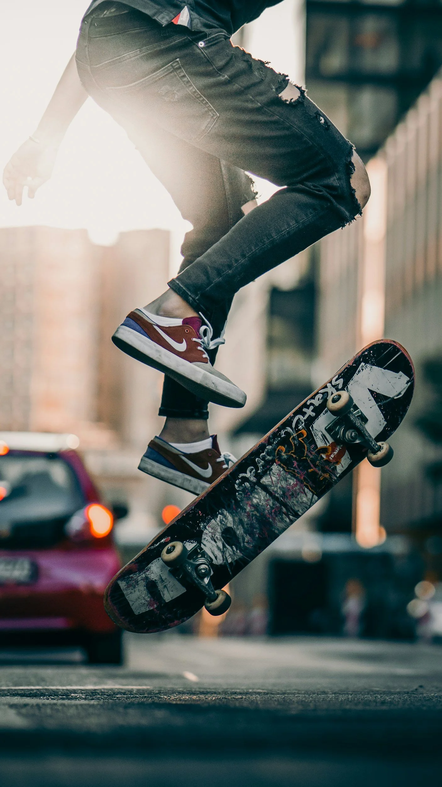 A person performing a skateboarding trick in mid-air on an urban street, wearing red and white Nike sneakers, ripped black jeans, and a dark top.