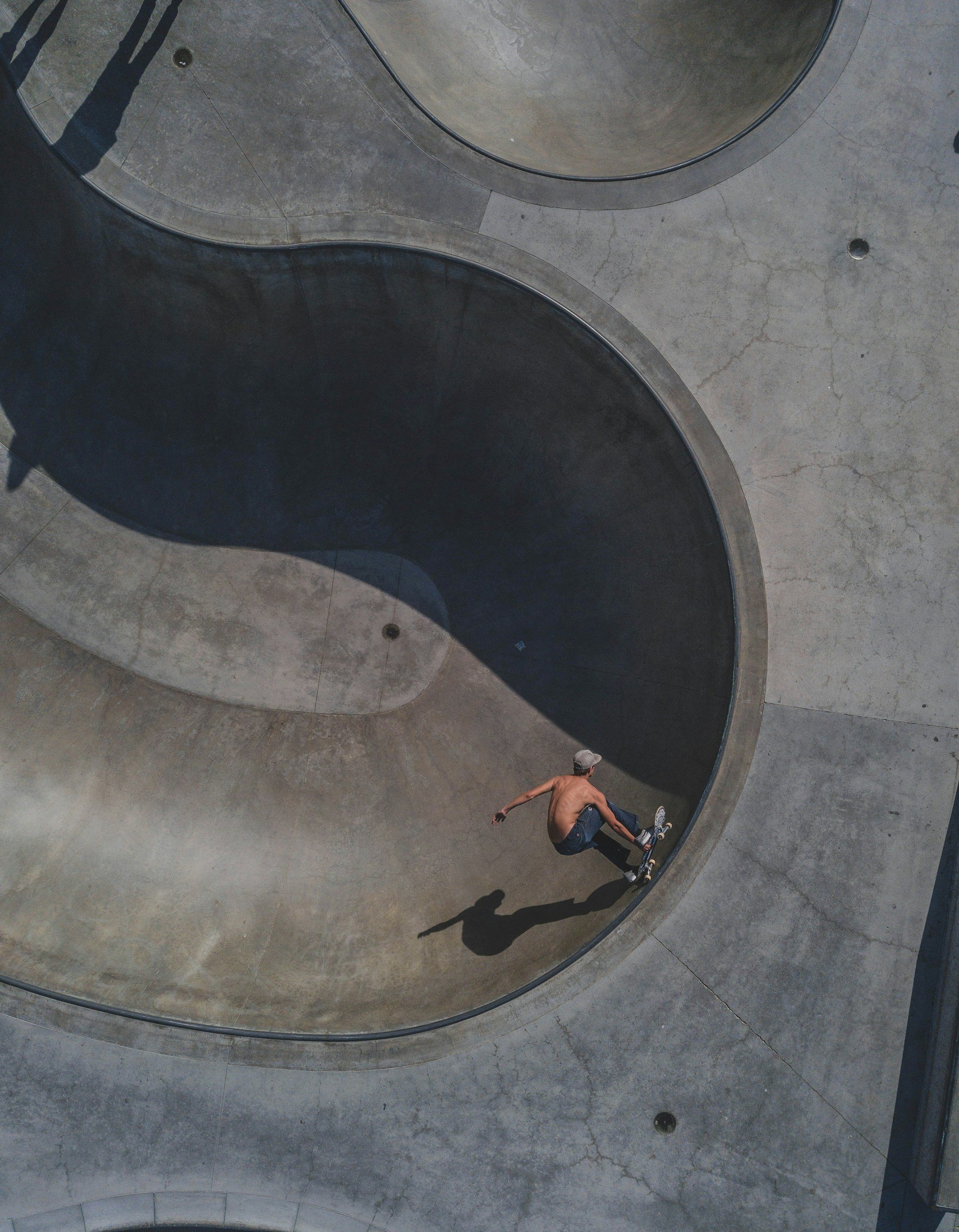 A shirtless skateboarder wearing a gray cap riding his skateboard in a concrete skatepark with curved bowls, casting a shadow on the ground.