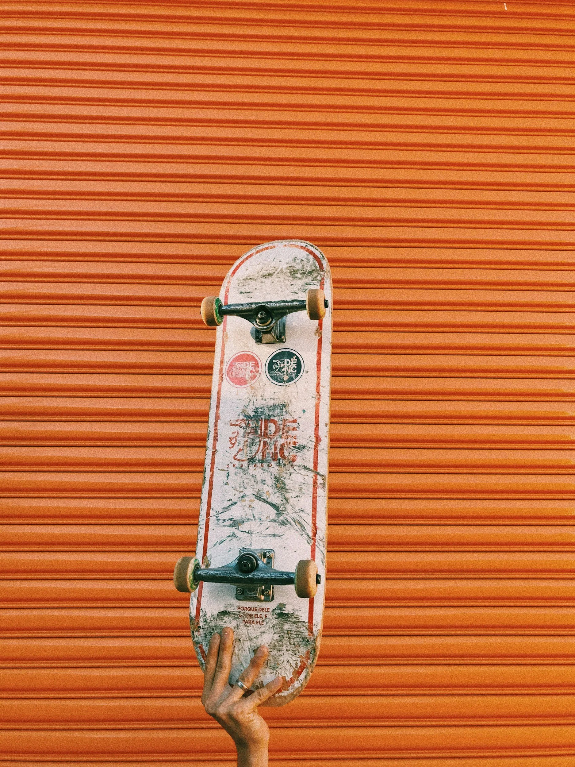A skateboard held by a hand against an orange corrugated metal wall.