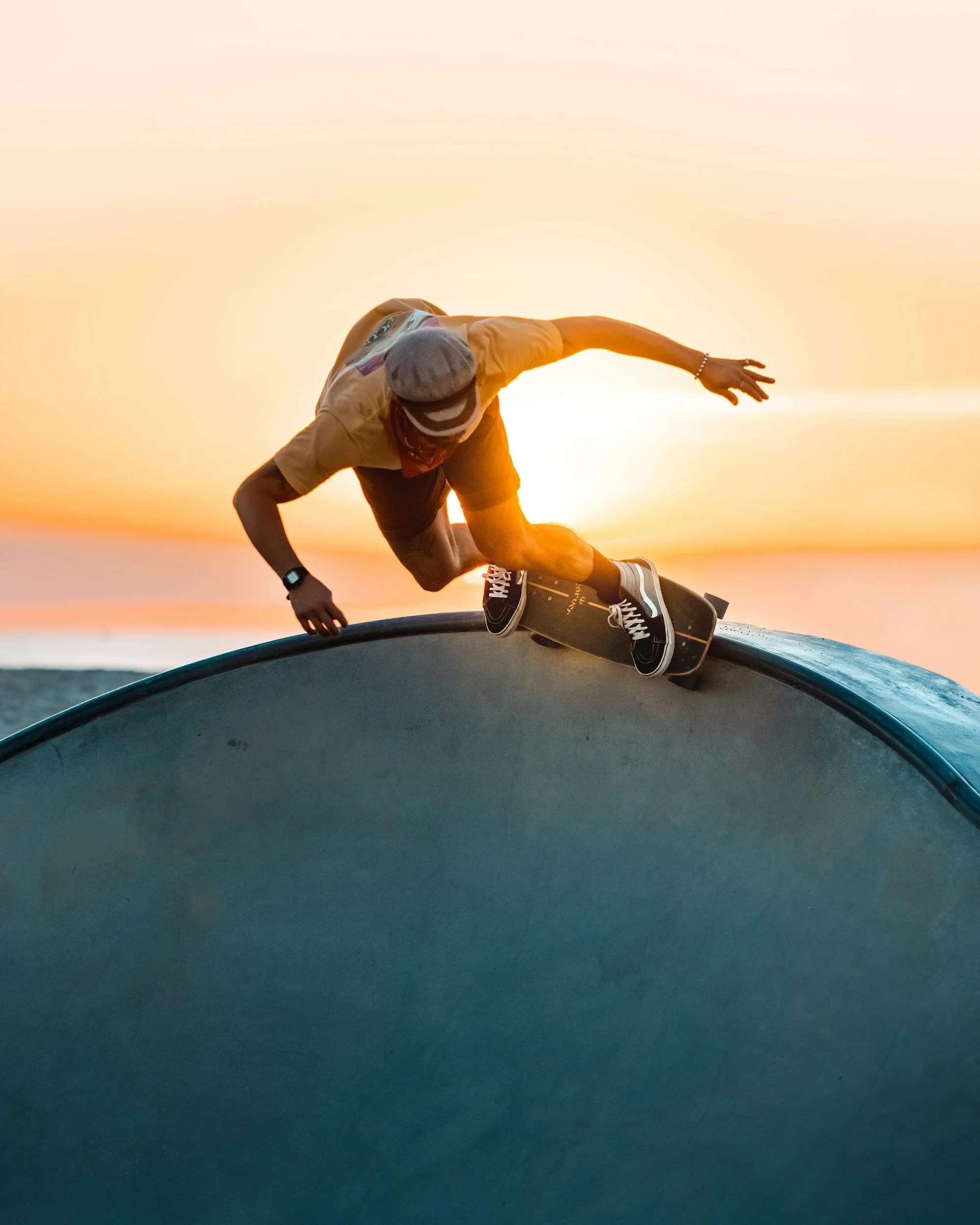 Skateboarder wearing a beanie cap and sunglasses performs a trick on a skateboard at sunset on a curved skate ramp.